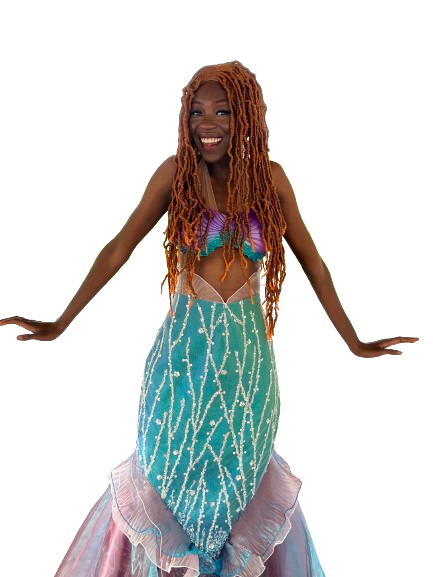 A woman in a colorful mermaid costume with a purple and blue top, a turquoise tail with white patterns, and a pink and white layered skirt, smiling with her arms outstretched.