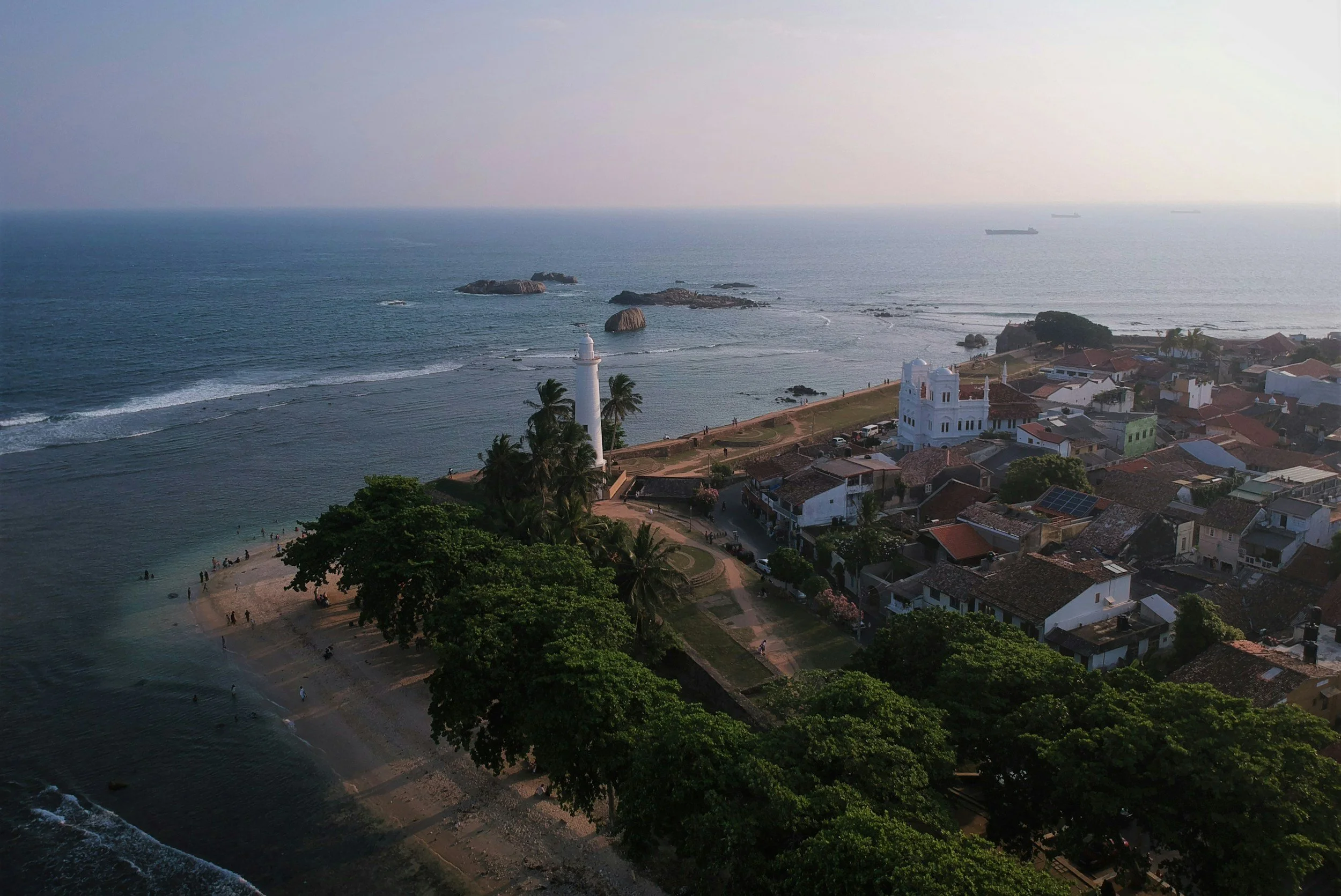Vista aérea de un pueblo costero con un faro grande y una playa con personas nadando, rocas en el mar, y casas tradicionales con techos de teja roja, rodeado de árboles verdes y vegetación.