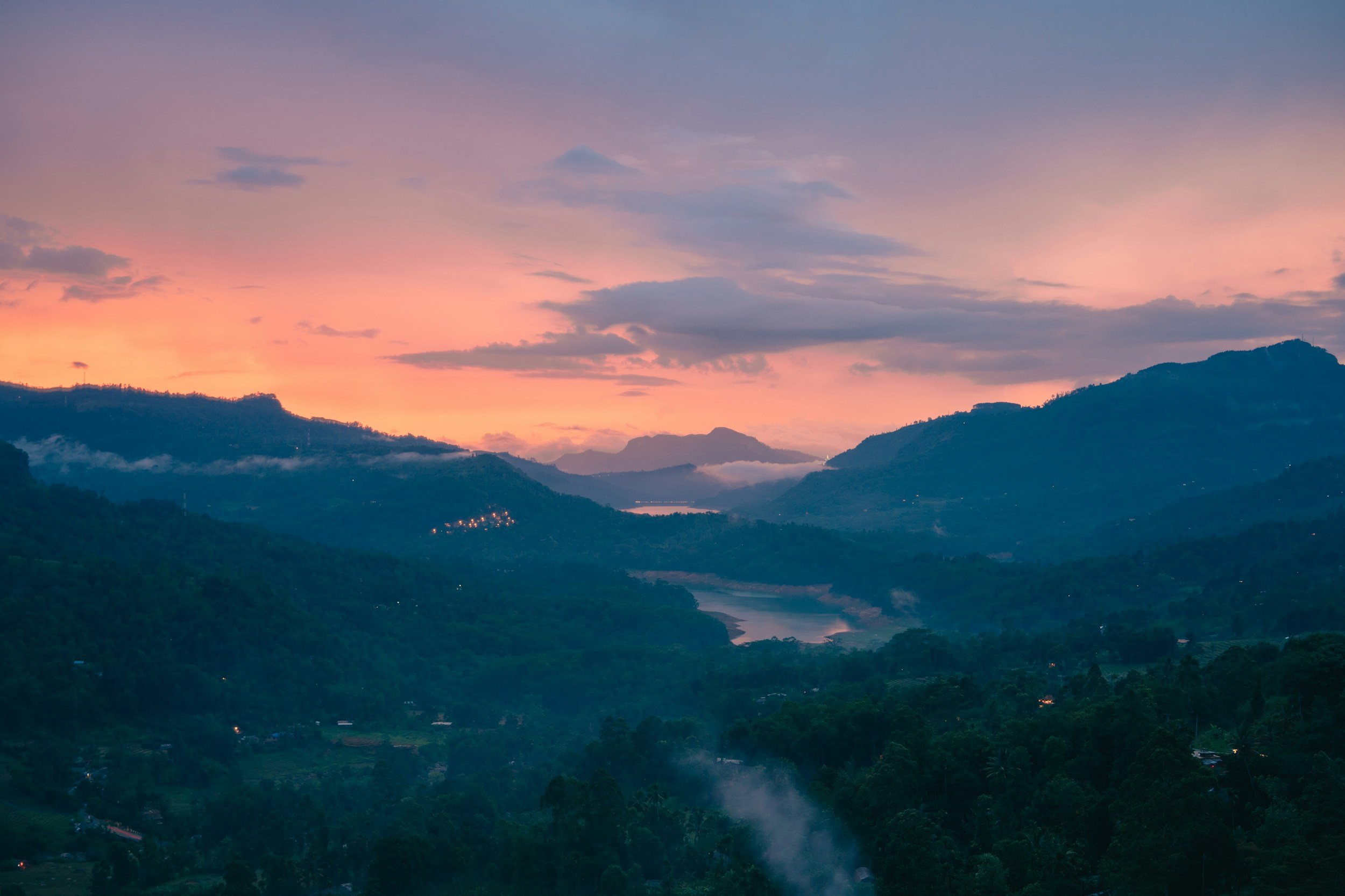 Paisaje montañoso al atardecer con cielo color rosa y azul, con una corriente de agua que atraviesa el valle cubierto de vegetación y algunas nubes en la montaña.