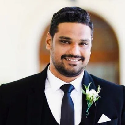 Hombre sonriendo con traje formal y flor en el ojal, en un fondo difuso y cálido.