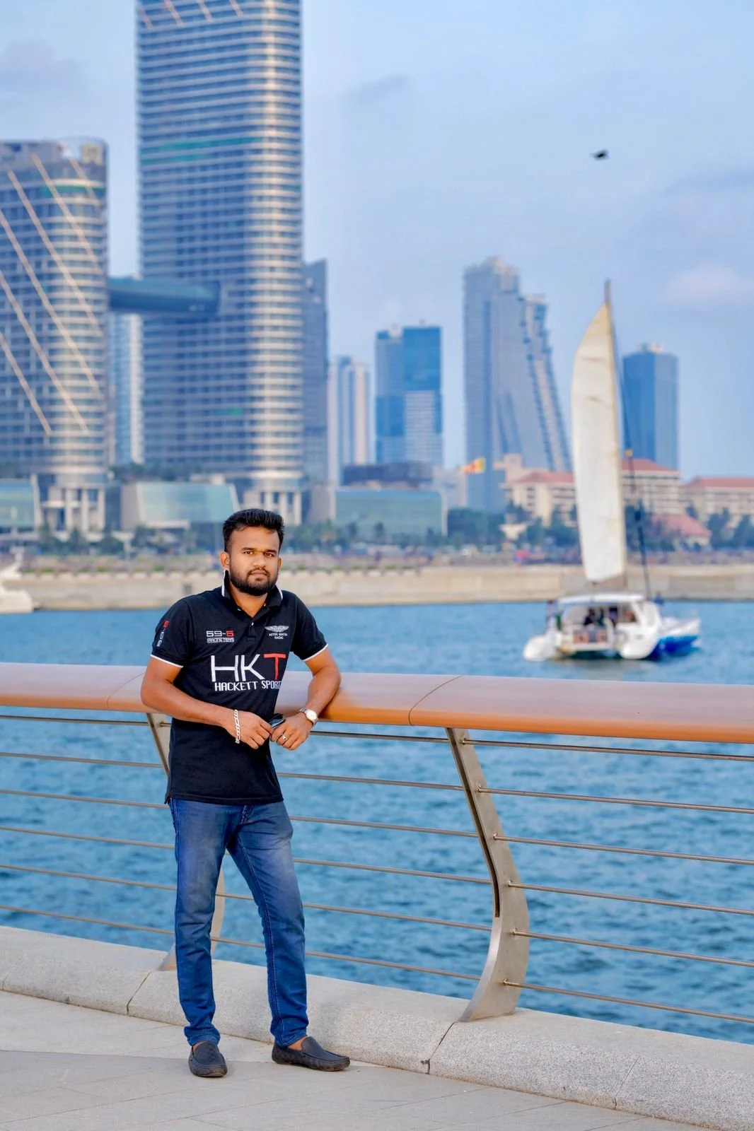Un hombre de futbolín con musculatura, pantalones cortos y camiseta negra, de pie junto a un paseo junto al agua en una ciudad moderna con rascacielos.