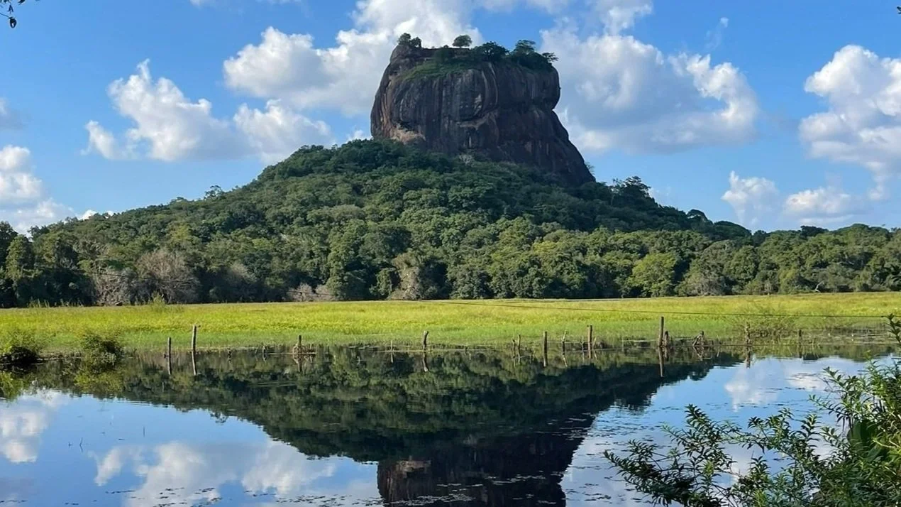 Una roca grande y aislada en forma de meseta en la cima de una colina cubierta de árboles, reflejada en un río tranquilo, con nubes en el cielo azul.