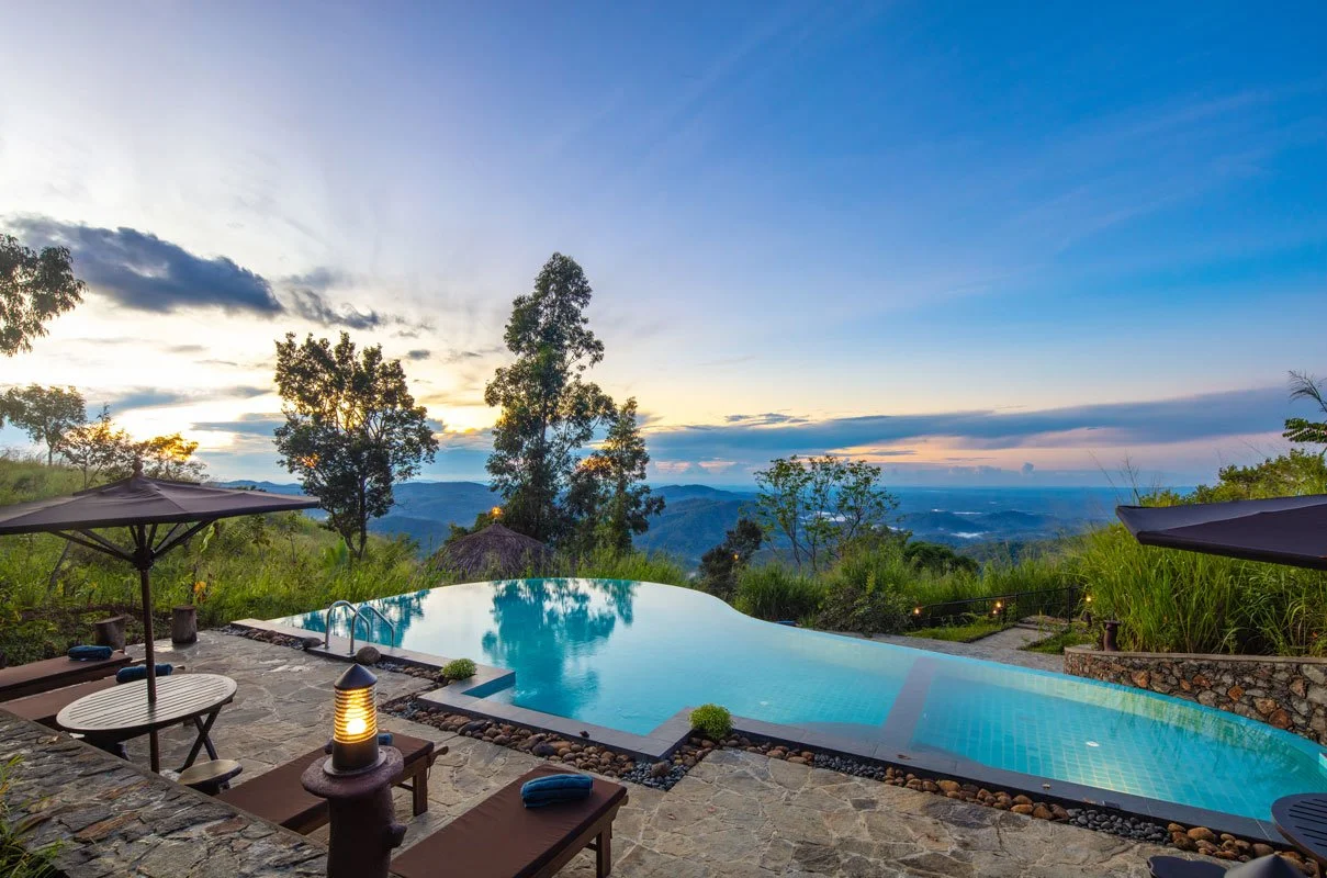 Vista de una piscina en un entorno natural con árboles, montañas y un atardecer, con sillones y sombrillas alrededor.