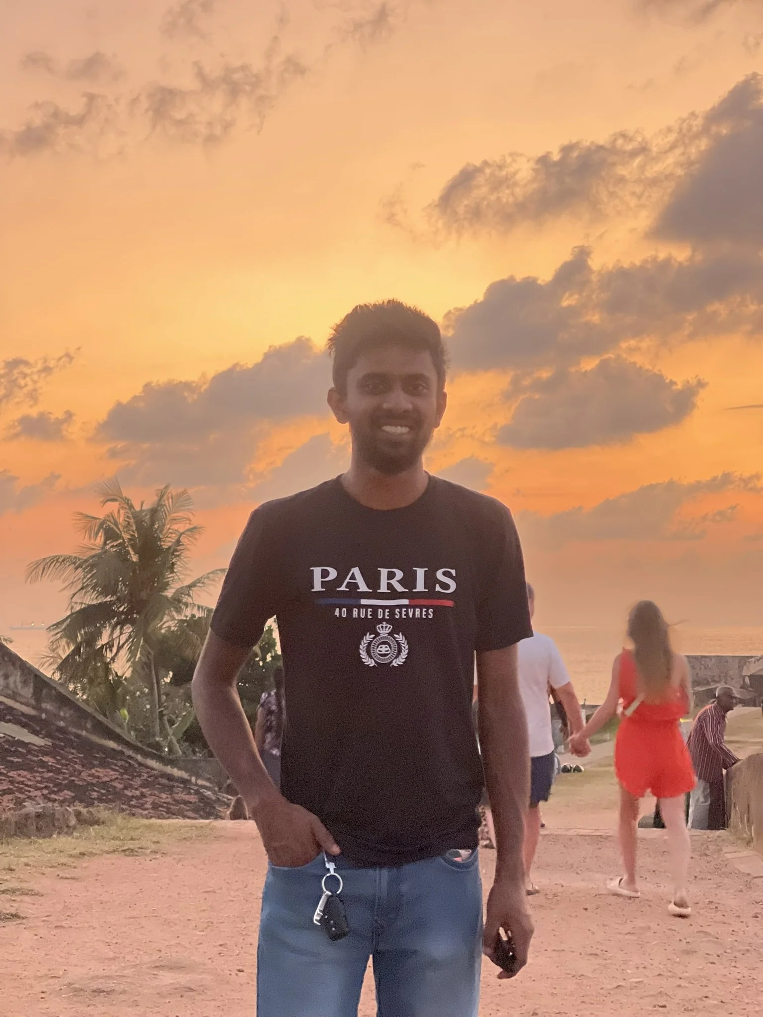 Joven sonriendo en un lugar al atardecer, con cielo nublado y árboles de palma, y otras personas en el fondo caminando.