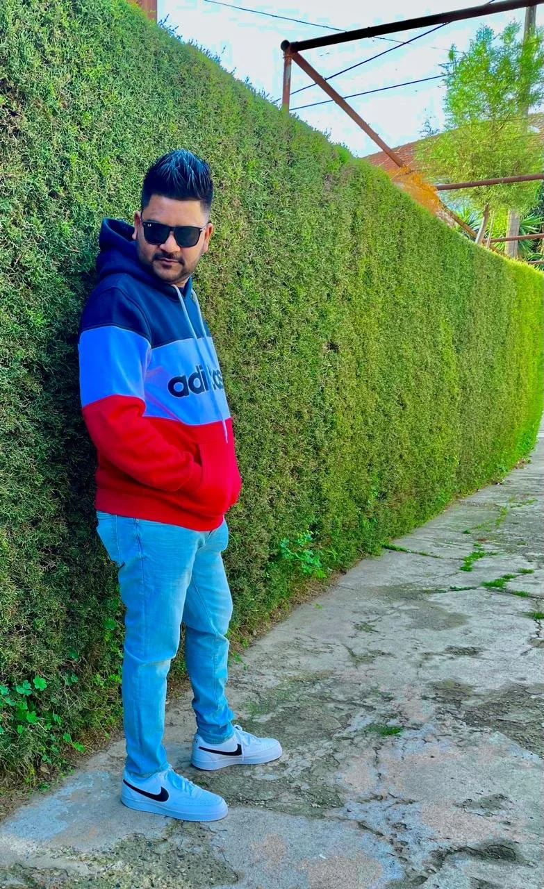 Un hombre con gafas oscuras, sudadera con capucha en colores azul, rojo y negro, jeans claros y zapatillas blancas, apoyado contra una cerca de arbustos verdes.