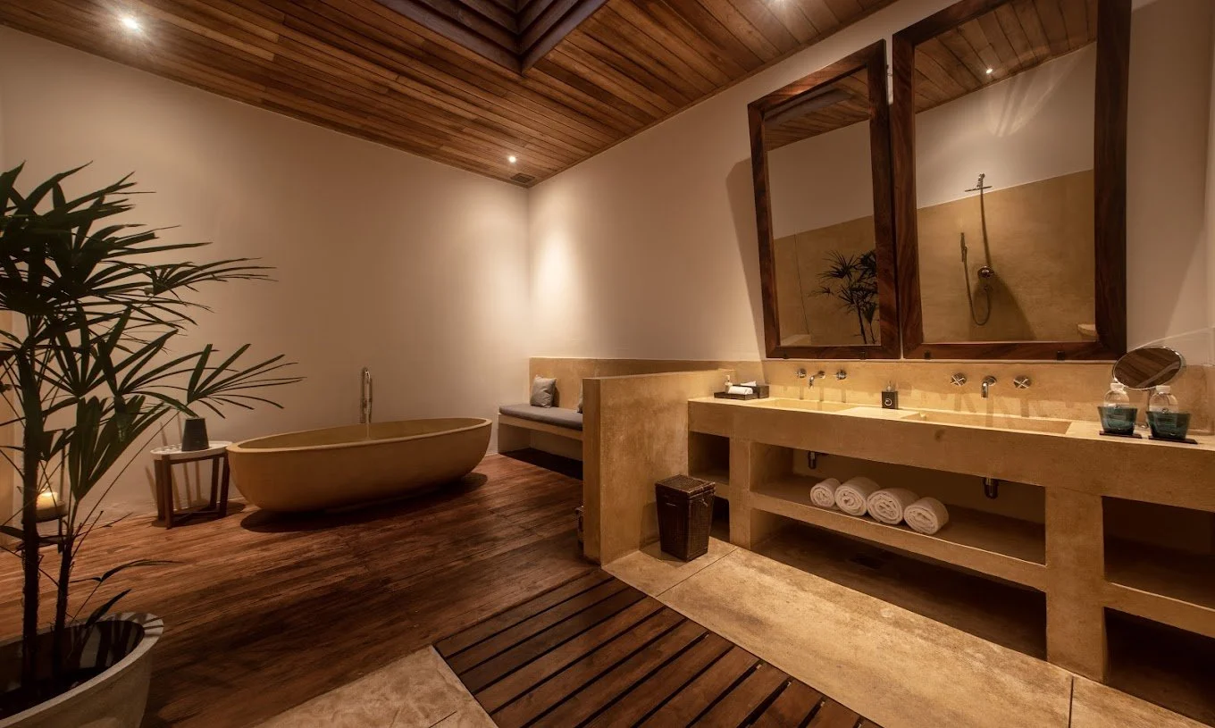 Un baño de spa con bañera de piedra, lavabo doble con espejo, piso de madera y plantas decorativas.