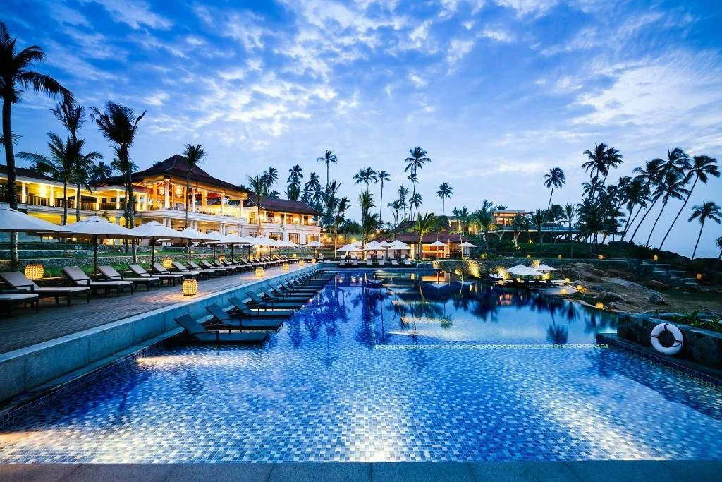 Piscina en un hotel con sillas y sombrillas, rodeada de palmeras y un edificio de lujo, en una escena nocturna en un destino tropical.