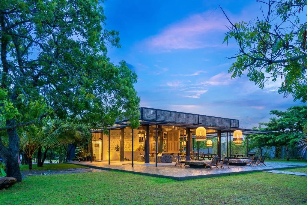 Casa moderna de un solo piso con terraza y muebles de exterior en medio de un jardín con árboles, al atardecer.