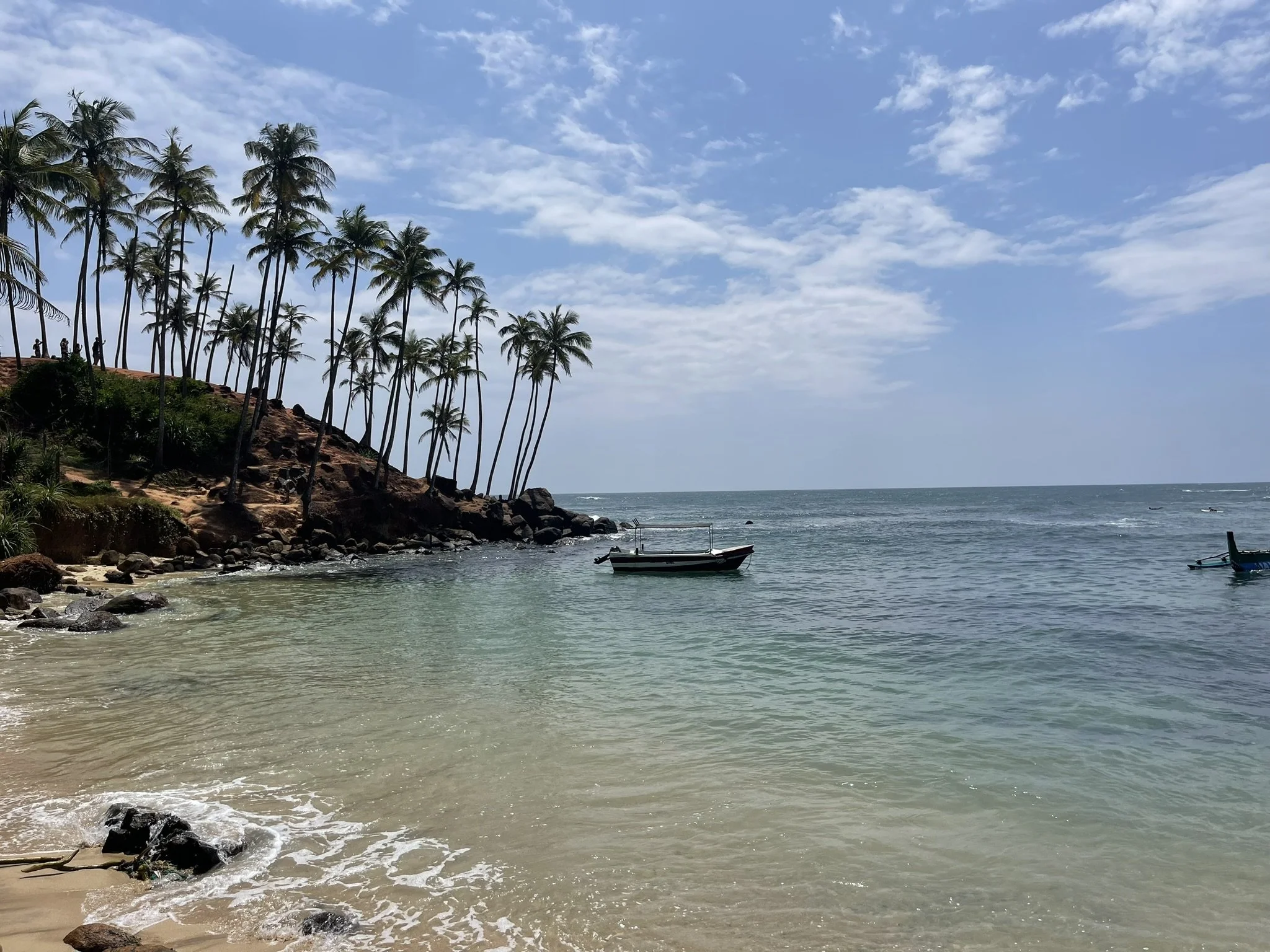 Vista de playa con palmeras en la orilla, varias embarcaciones en el agua y cielo parcialmente nublado.