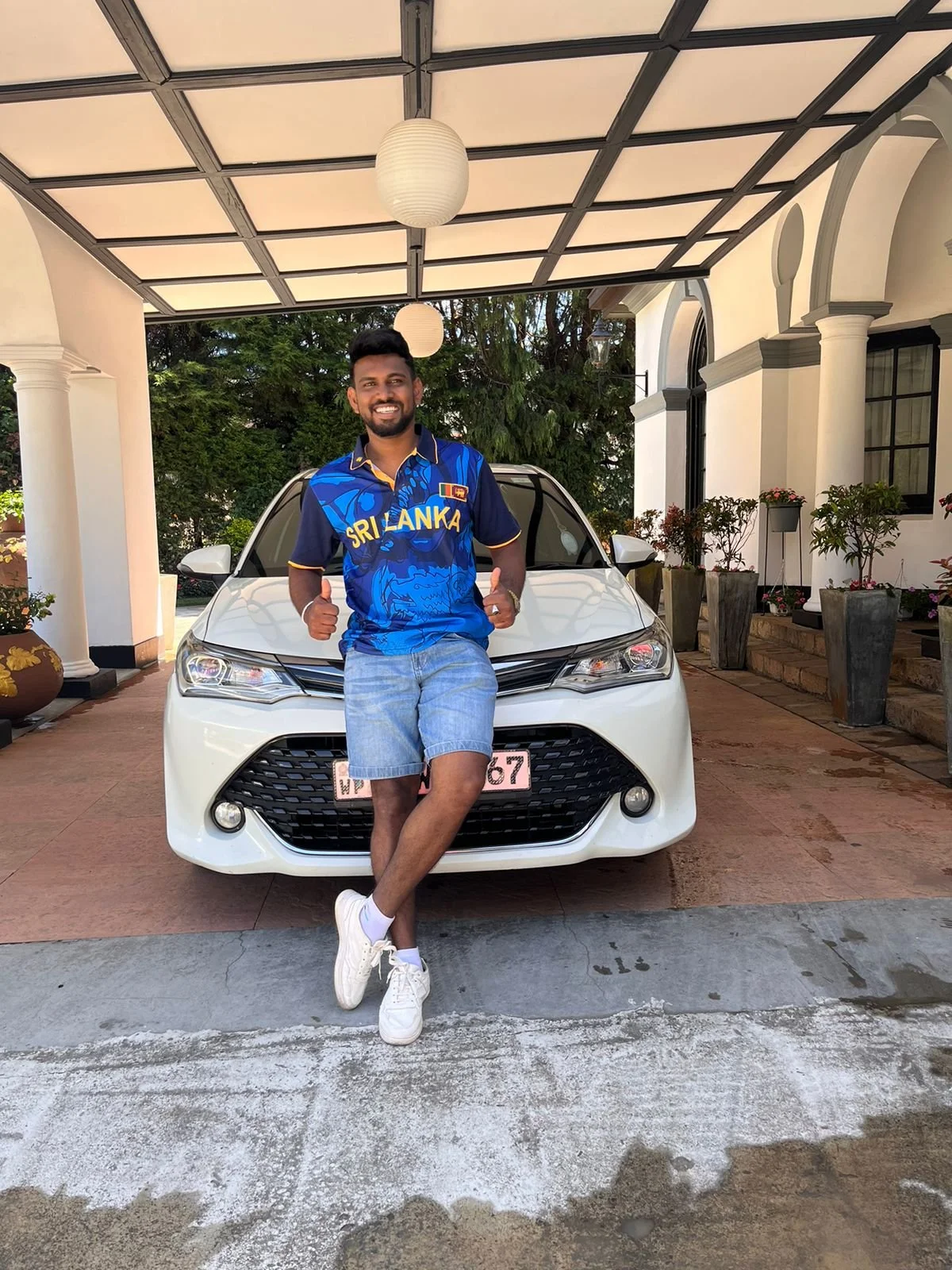 Hombre joven con camiseta de Sri Lanka, pantalones cortos y zapatillas blancas, sonriendo y haciendo pulgares hacia arriba en frente de un coche blanco en un patio con plantas y una estructura con cubierto.
