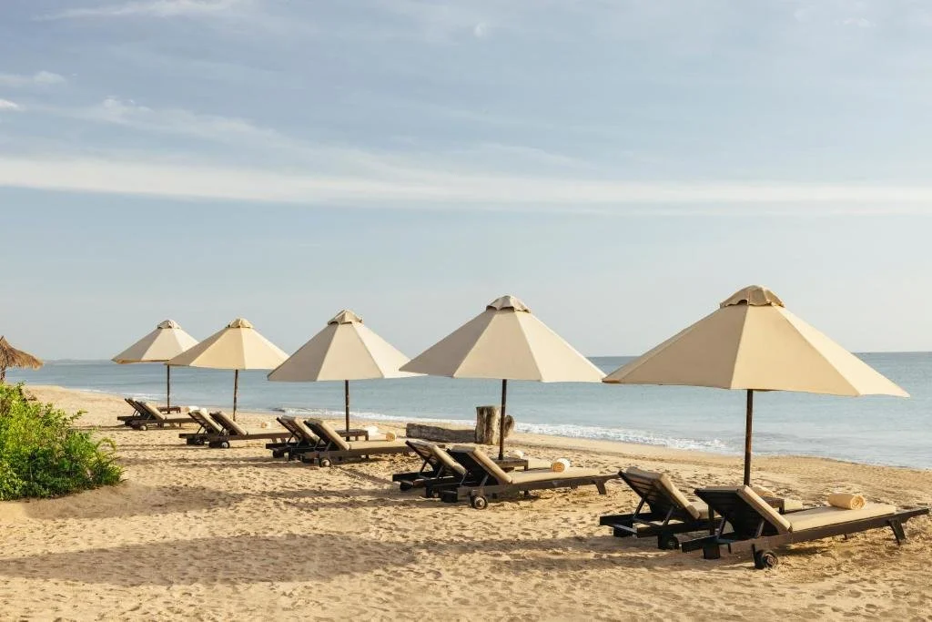 Una fila de tumbonas y sombrillas beige en una playa arenosa con el mar y un cielo parcialmente nublado de fondo.