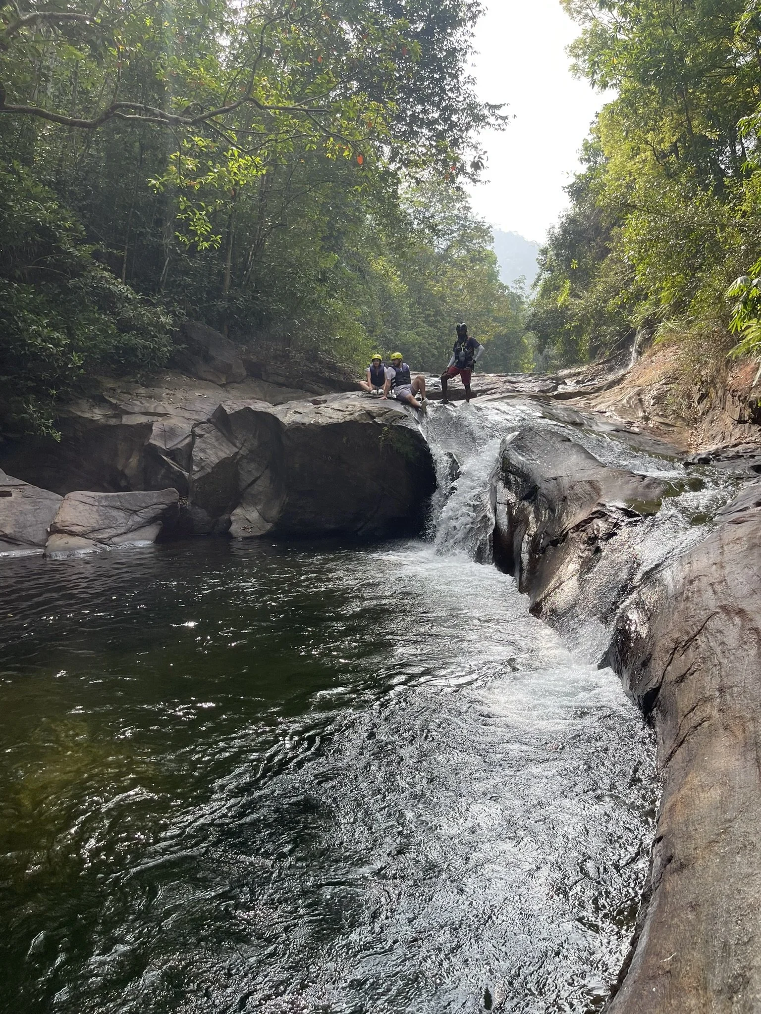 Tres personas en una zona de río con agua y rocas en un entorno de selva, una de ellas de pie y las otras dos sentadas en una roca. 