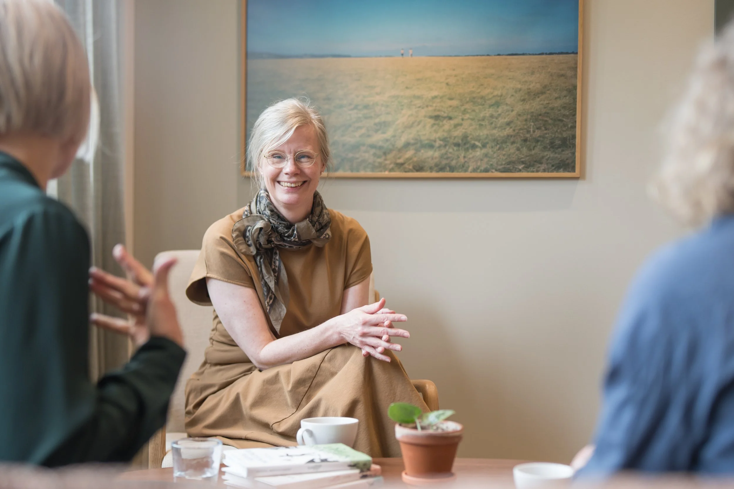 En gruppe mennesker i samtale, med en kvinne i midten som smilende snakker, i et rom med et landskapsbilde på veggen