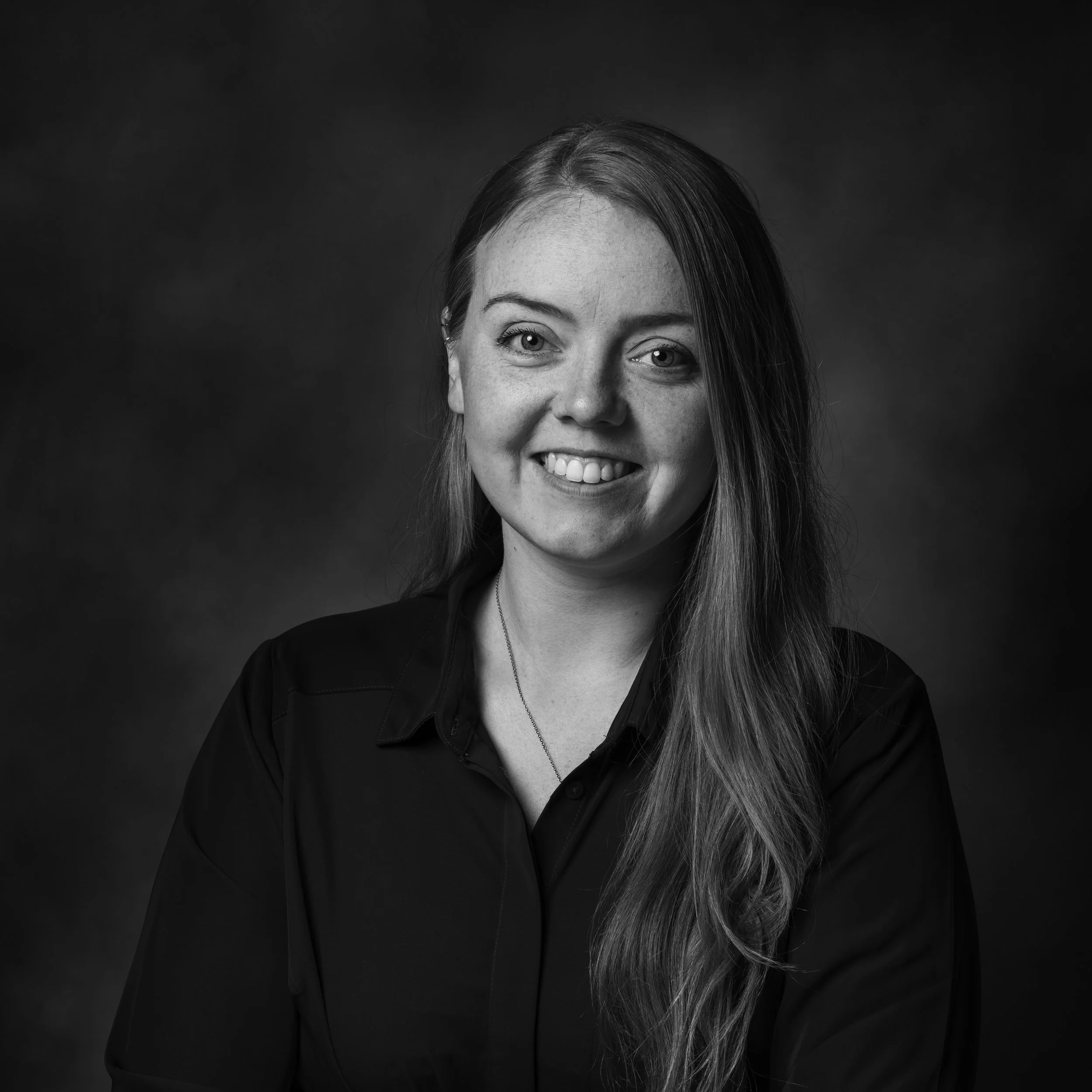 Sort-hvitt fotografi av en smilende kvinne med langt hår i svart skjorte, mot en mørk bakgrunn.