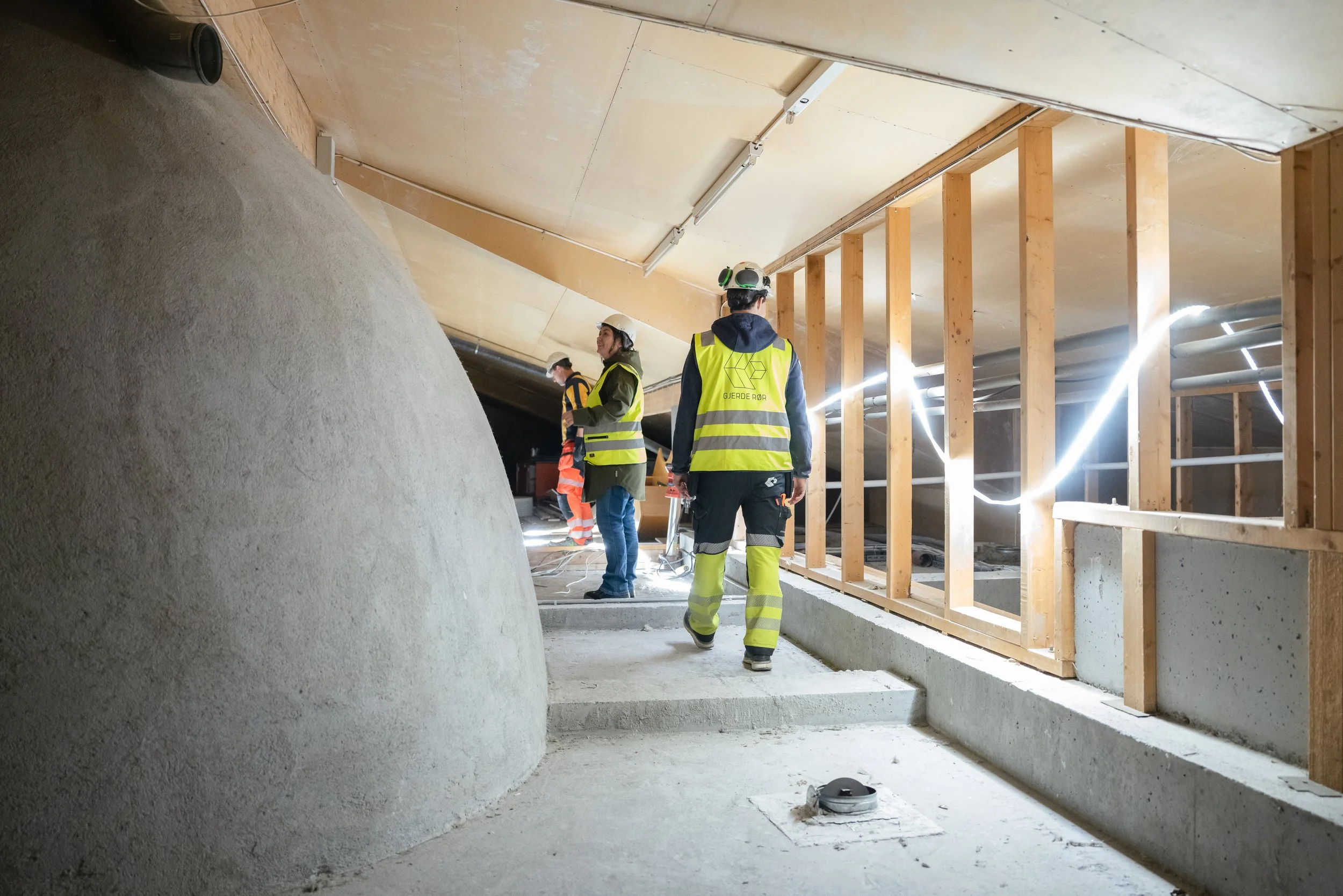 Bygningsarbeidere i sikkerhetsutstyr som jobber inne i en underjordisk konstruksjon, med treverk, betong og elektriske kabler rundt seg.