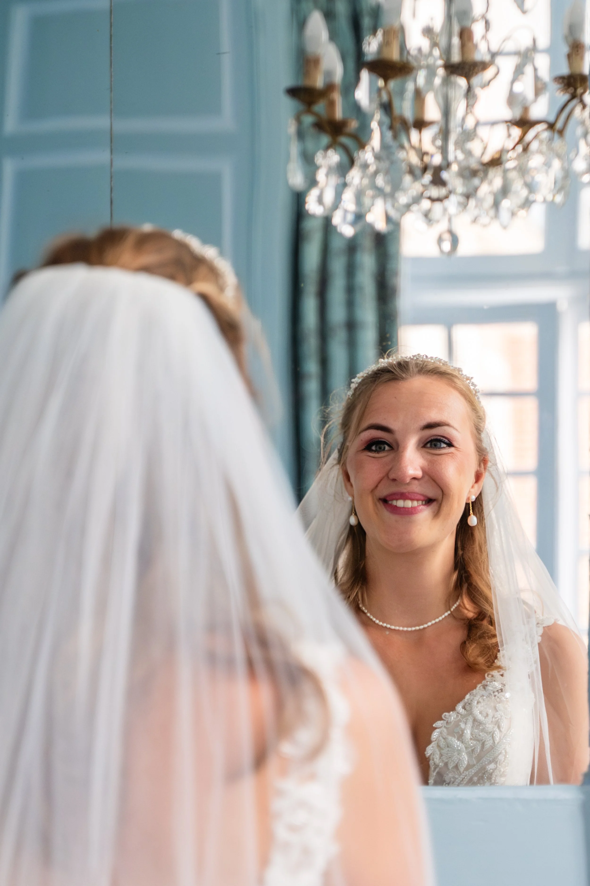 Vrouw in een trouwjurk die naar haar reflectie in de spiegel kijkt, met een glimlach, in een elegante kamer met een kristallen chandelier.