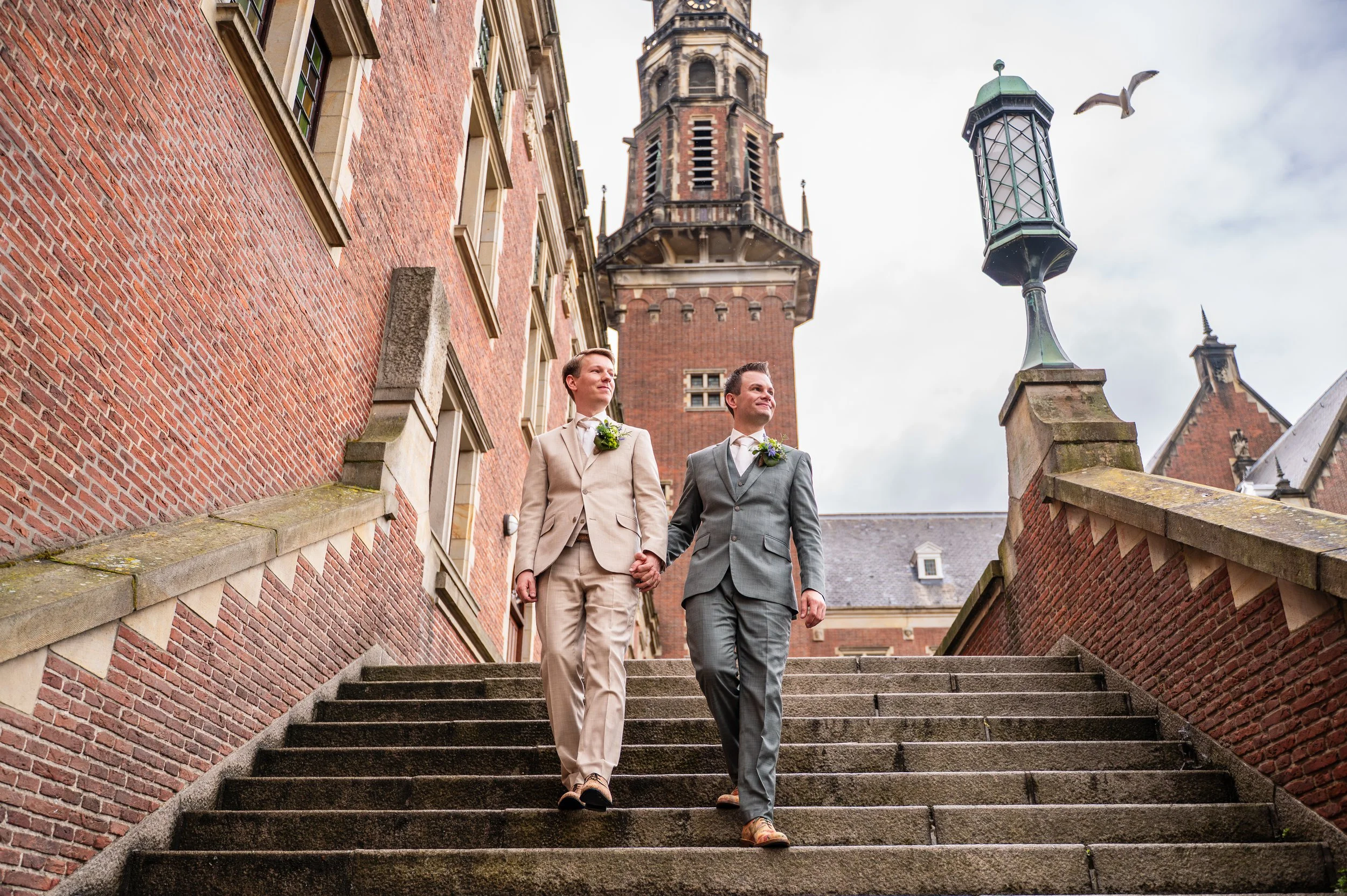 Twee mannen in huwelijkskleding lopen hand in hand naar beneden via een stenen trap voor een oude bakstenen gebouw met toren. Er vliegen vogels in de lucht.