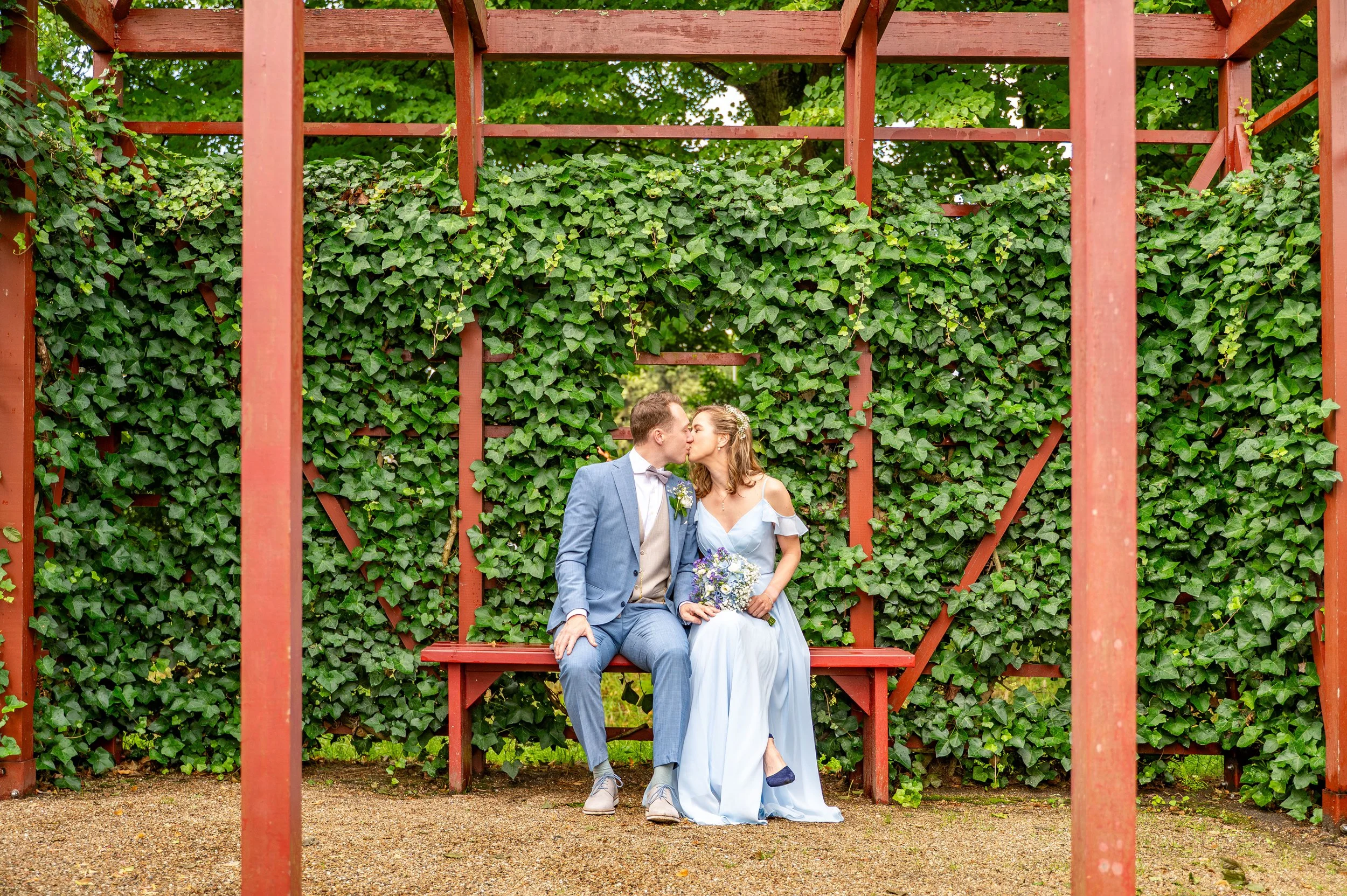 Een bruid en bruidegom zitten op een bankje in een tuin, kussen elkaar en delen een kus, omringd door een groene muur van bladeren.
