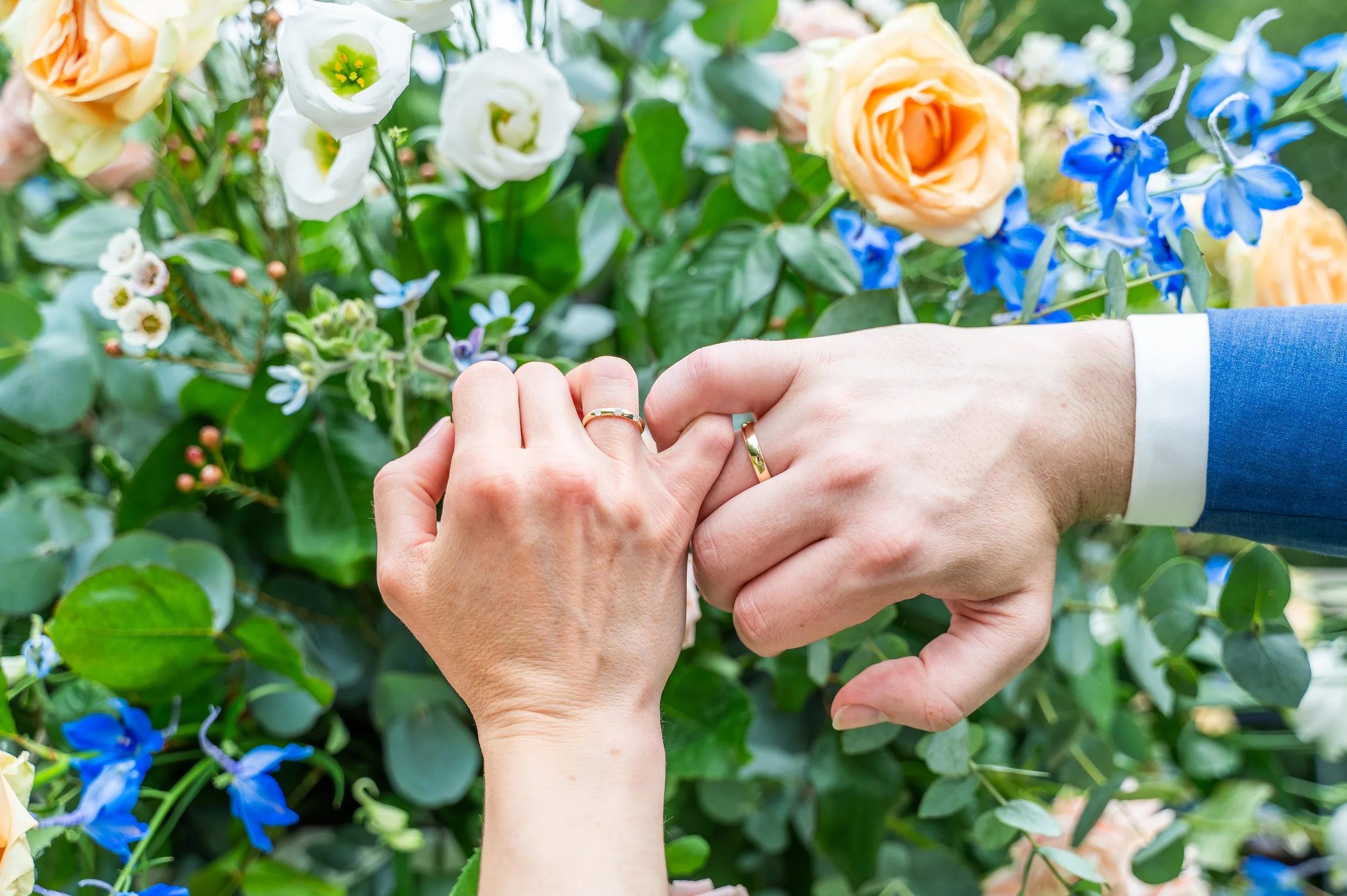 Twee personen met gepakt handen, beiden dragen ringen, tegen een achtergrond van bloemen.