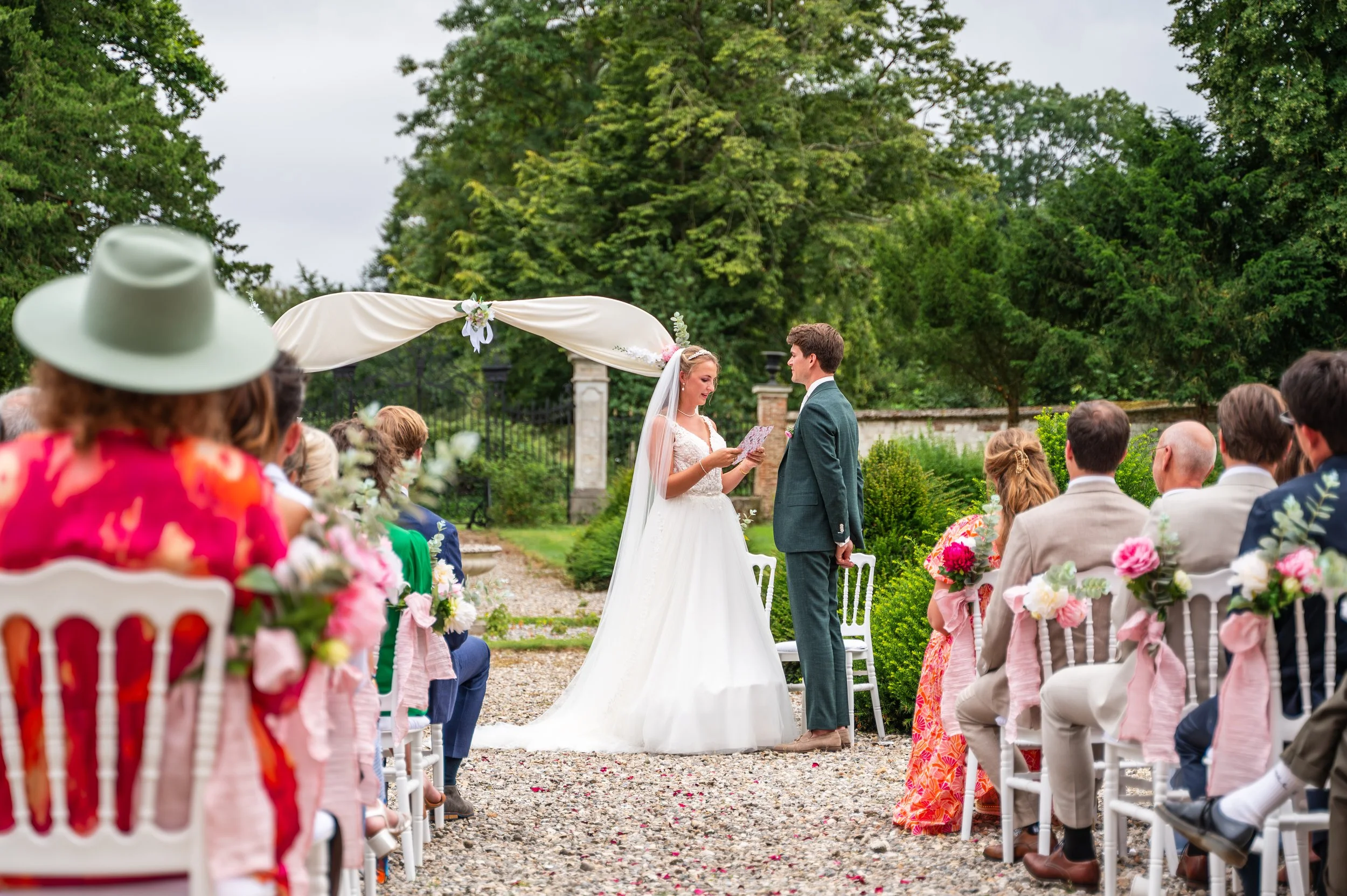 Bruidsgetrouwdepaar onder huwelijksboog tijdens ceremoni, omringd door zittende gasten op buitenlocatie met groene bomen en tuin.
