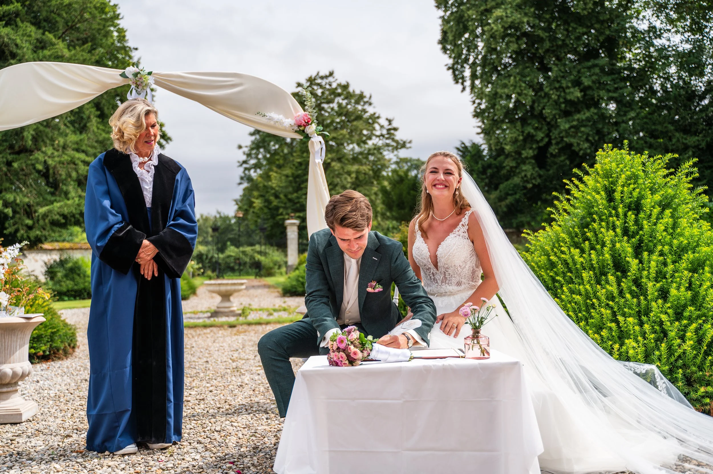 Bruidegom die ondertekenen het huwelijkscontract, terwijl de bruid lachend naast hem staat tijdens een buitenceremonie. Een officiëel staat ernaast, en er is een versierd bloemenboog en groene bomen op de achtergrond.