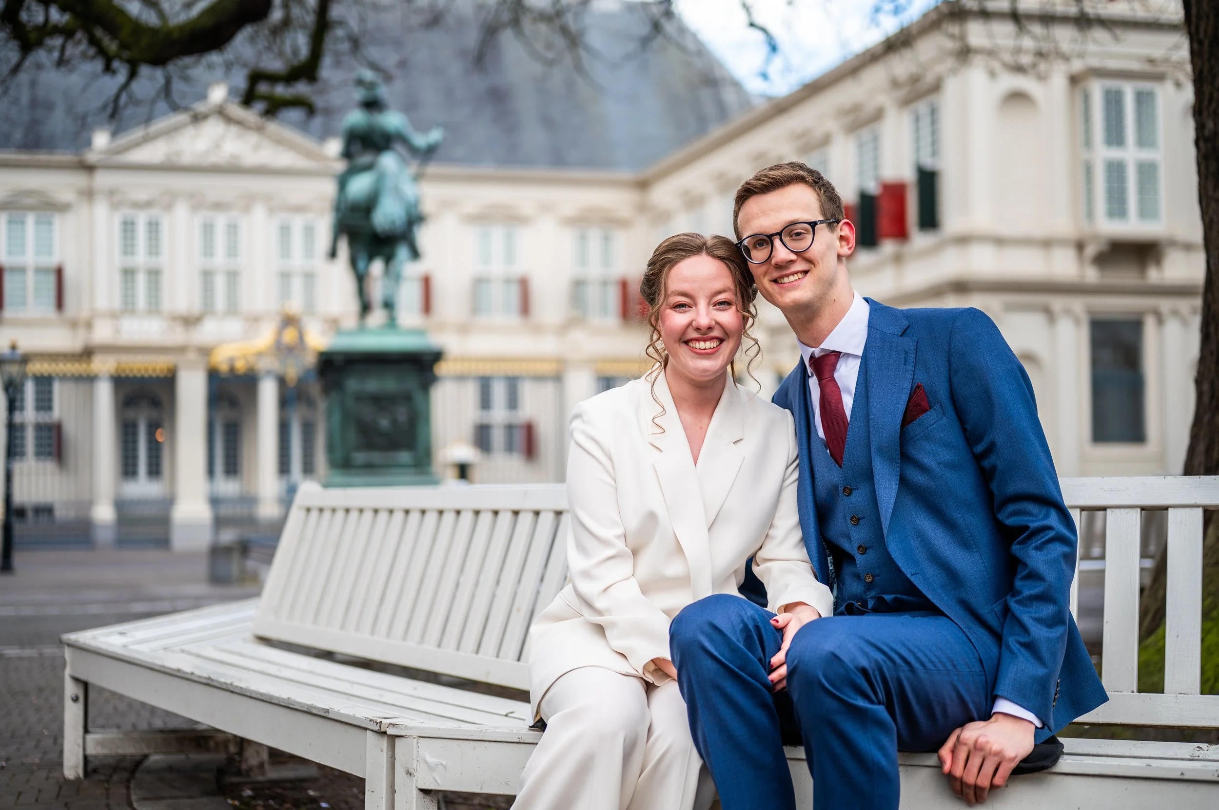Koppeltje zit op een witte bank in een park, lachend en gekleed voor een formeel evenement, met een standbeeld en een groot gebouw op de achtergrond.