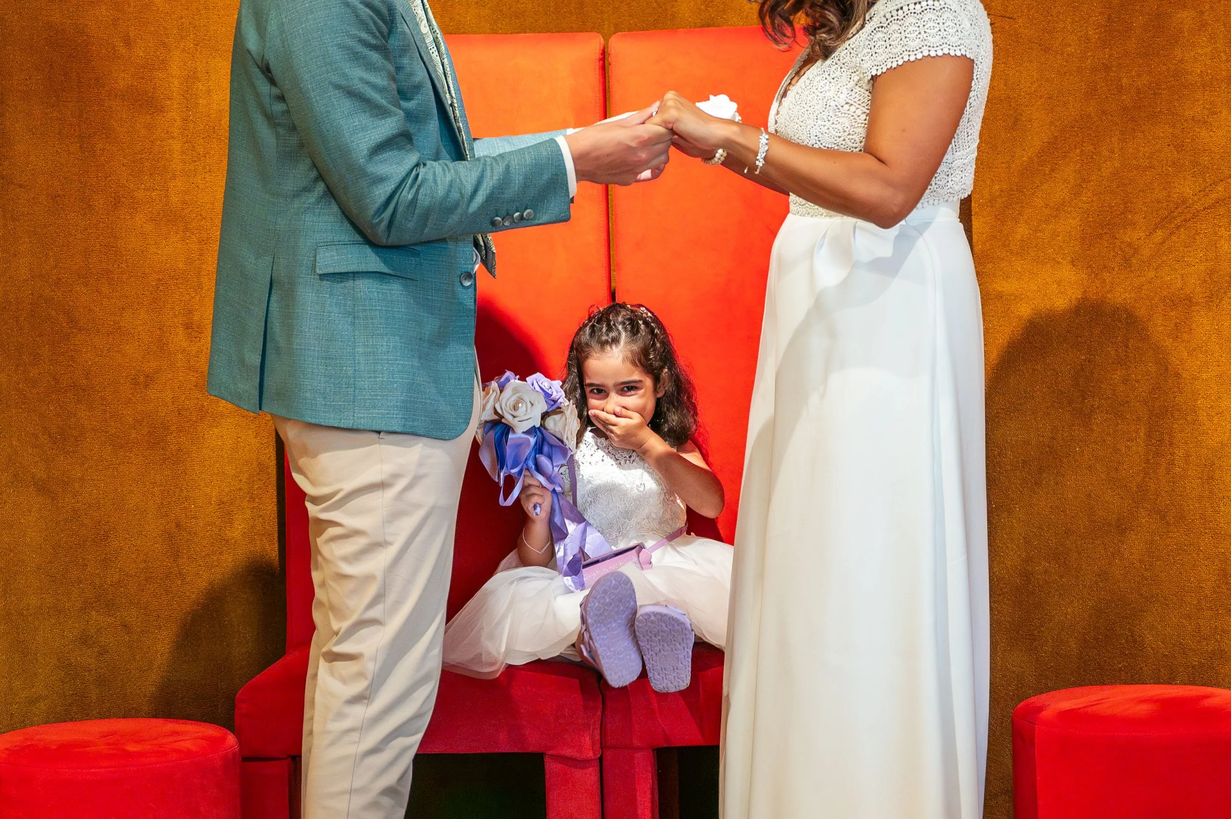 Een bruid en bruidegom houden handen vast tijdens een huwelijksceremonie, terwijl een jong meisje op een rode bank zit en een bos bloemen vasthoudt, met een verraste blik op haar gezicht.