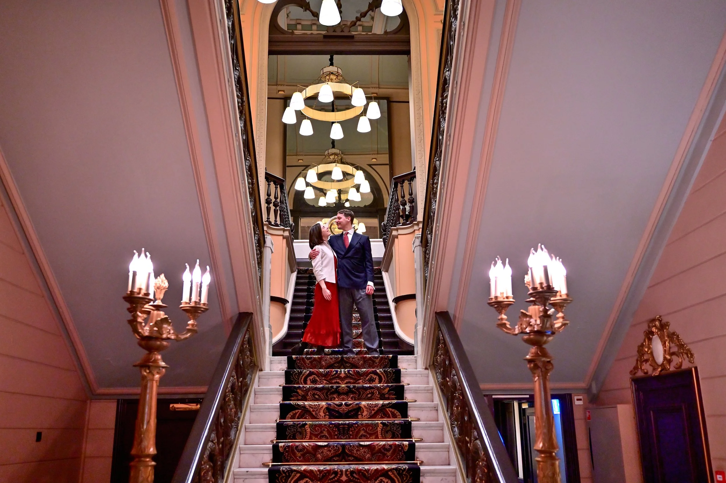 Een man en een vrouw staan samen op een trap in een elegant en klassiek interieur, omringd door kroonluchters en candelières, en kijken elkaar liefdevol aan.