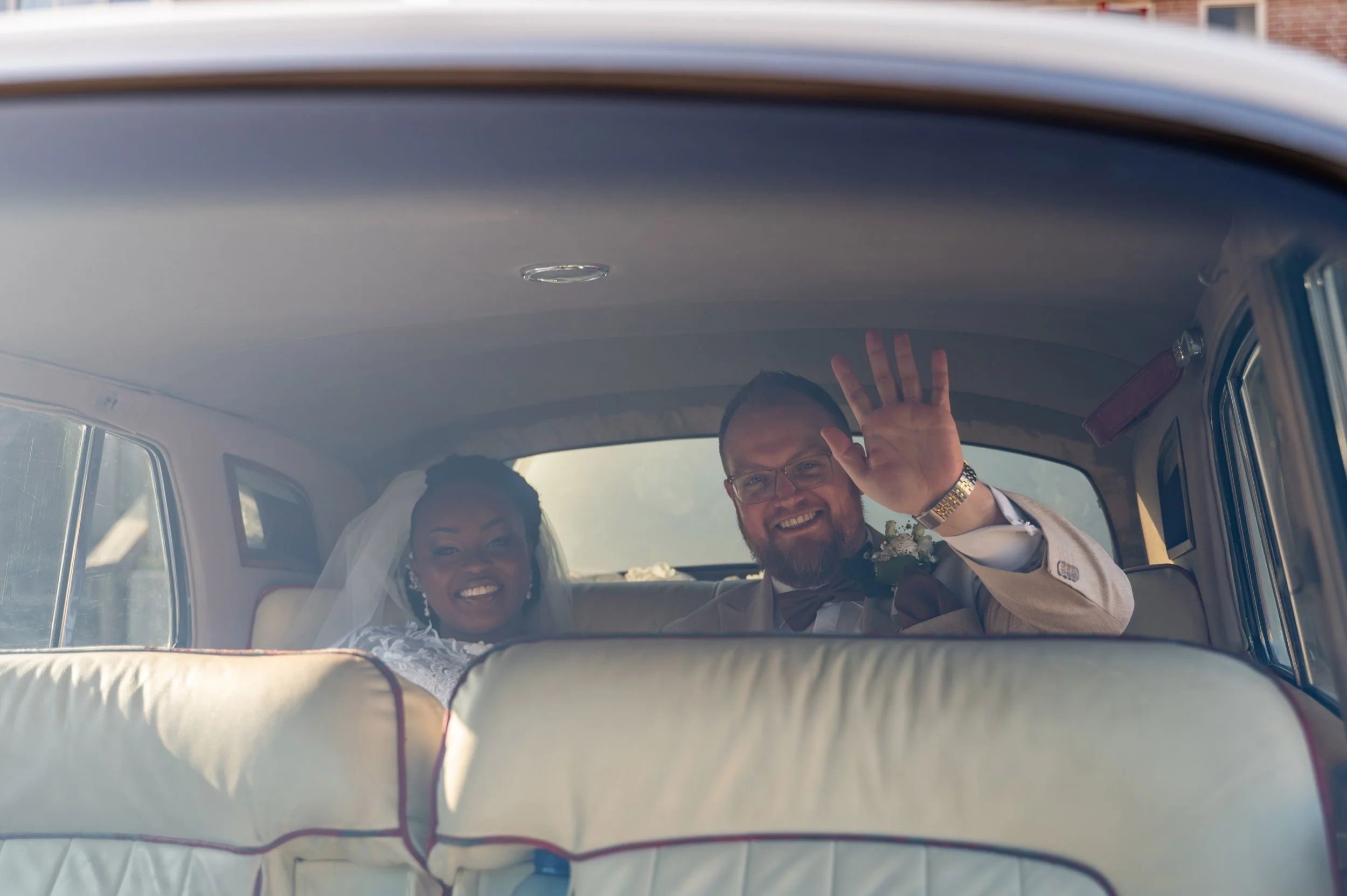 Een huwelijkspaar in de achterbak van een vintage auto, lachend en waving naar de camera.