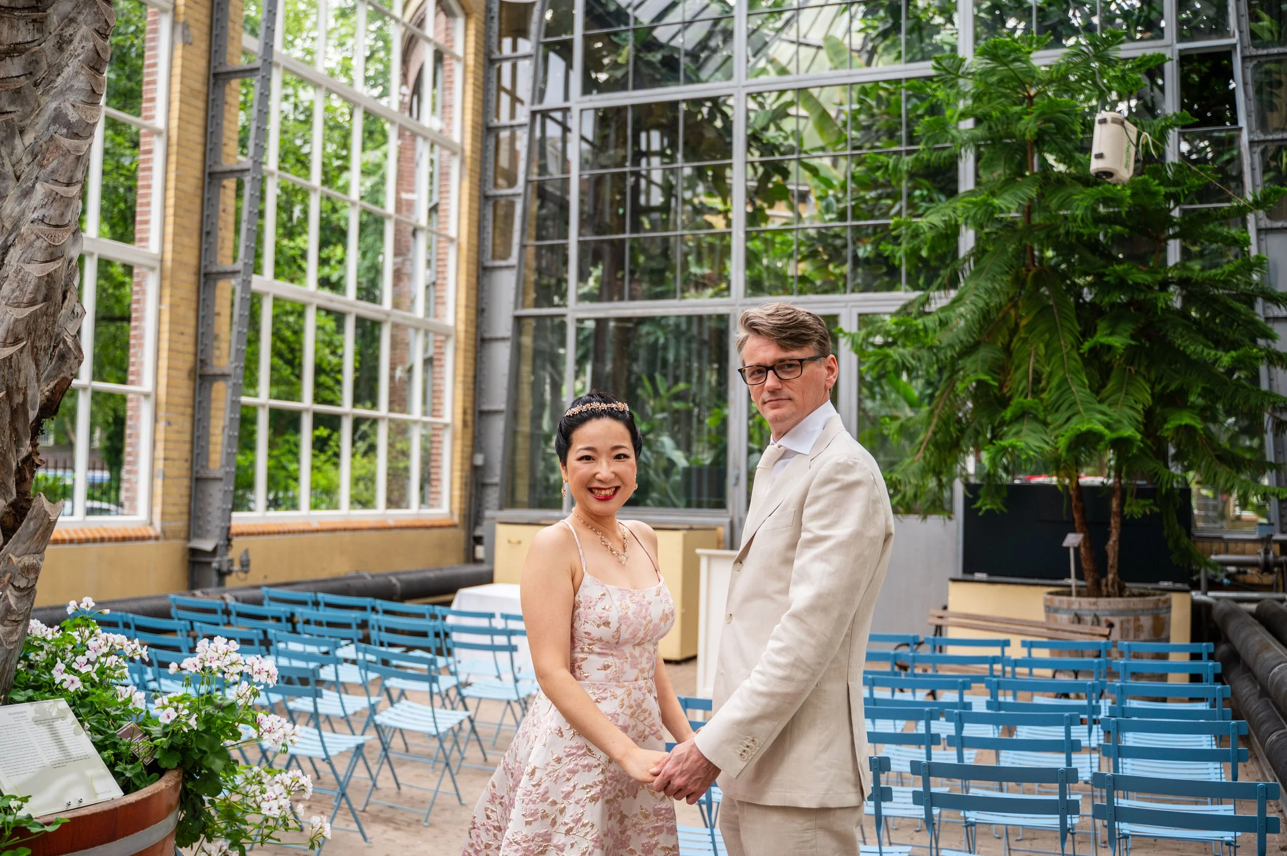 Een getrouwd paar houdt handen vast in een serre met veel groen en blauwe stoelen, de vrouw draagt een bloemachtig jurk en de man een lichtpak.
