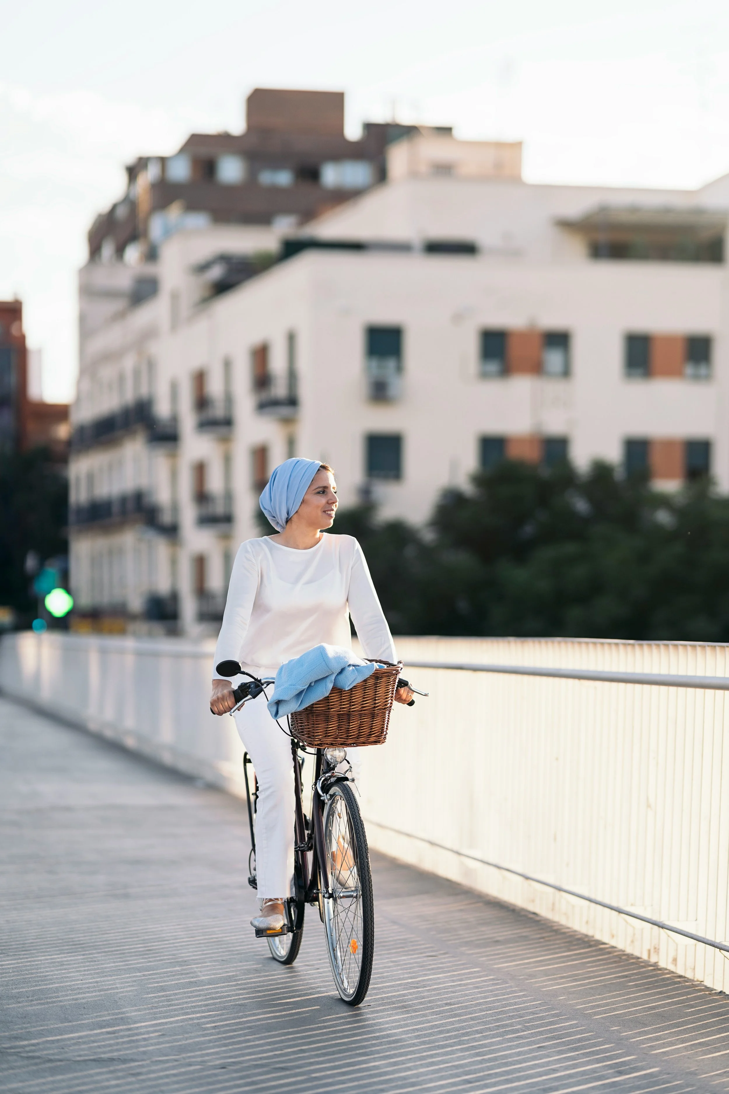 Femme souriante portant un hijab blanc, roulant à vélo avec panier en osier contenant un vêtement bleu, sur un pont urbain en fin d'après-midi.