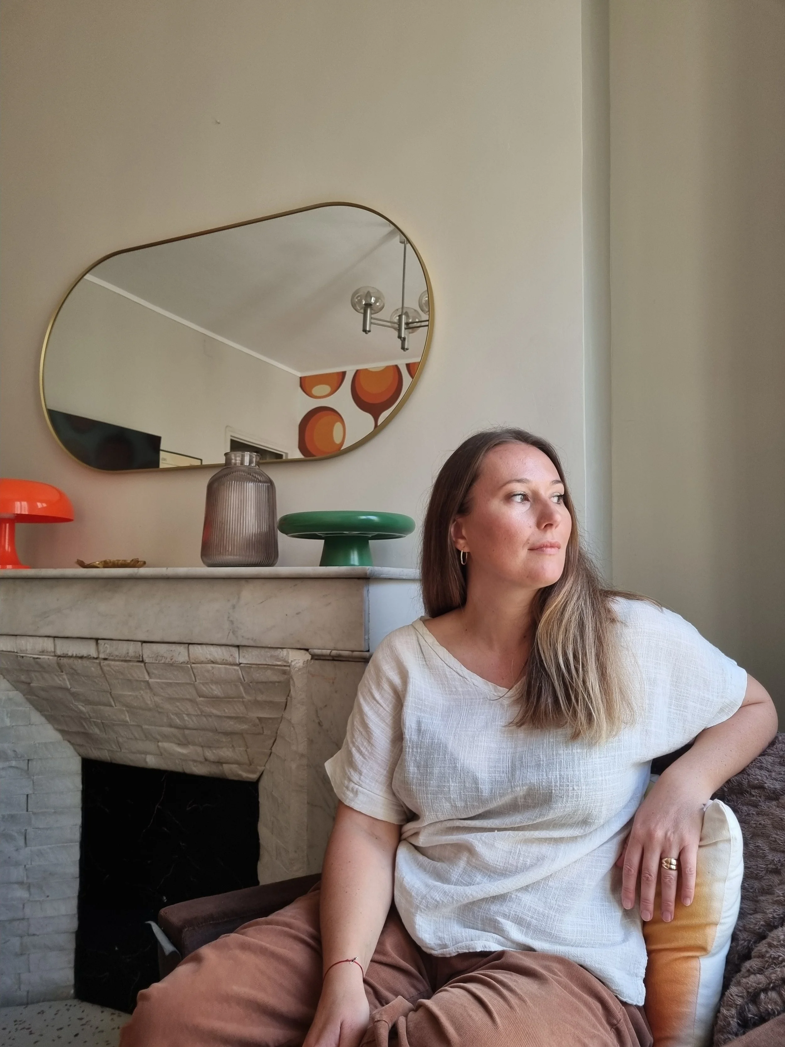 Une femme aux cheveux longs porte un t-shirt blanc et des pantalons marron. Elle est assise sur un canapé en cuir, à côté d'un coussin. Elle regarde vers la droite de l'image, dans un intérieur avec un miroir ovale au mur, et un décor rétro avec des cercles orange et marron.