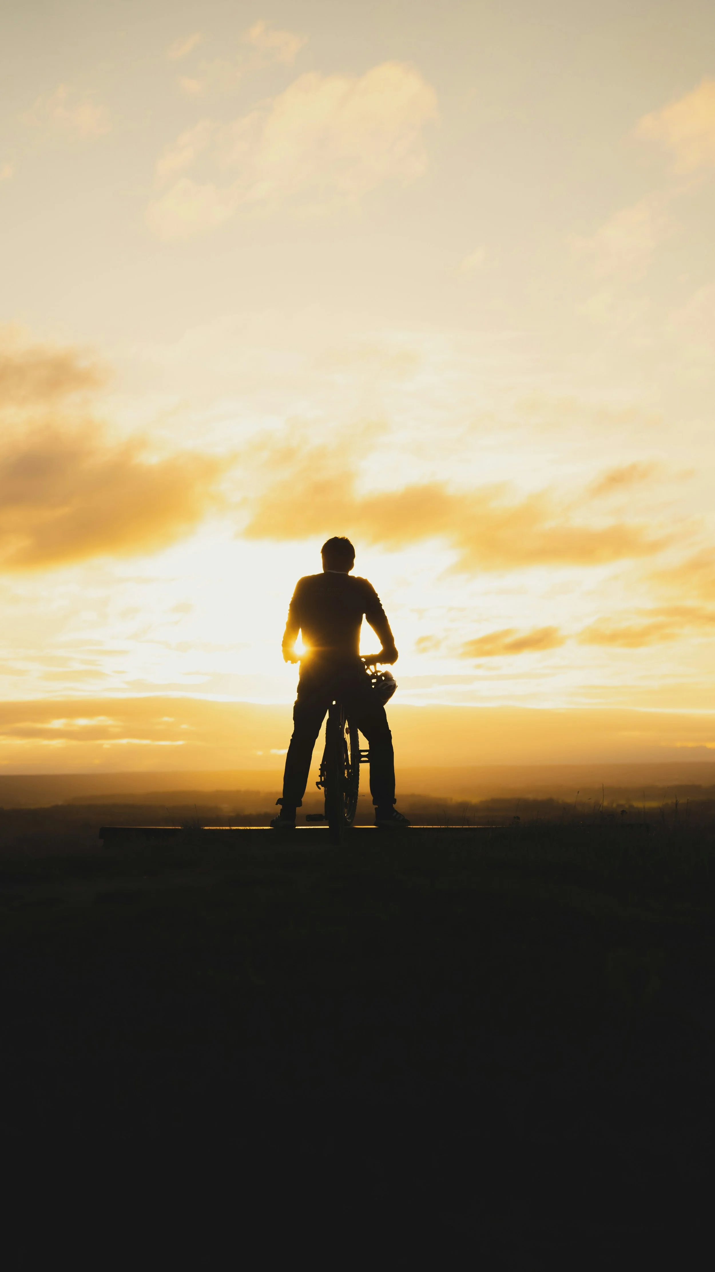 Une personne debout avec un vélo, en silhouette, regardant un coucher de soleil sur un horizon dégagé.