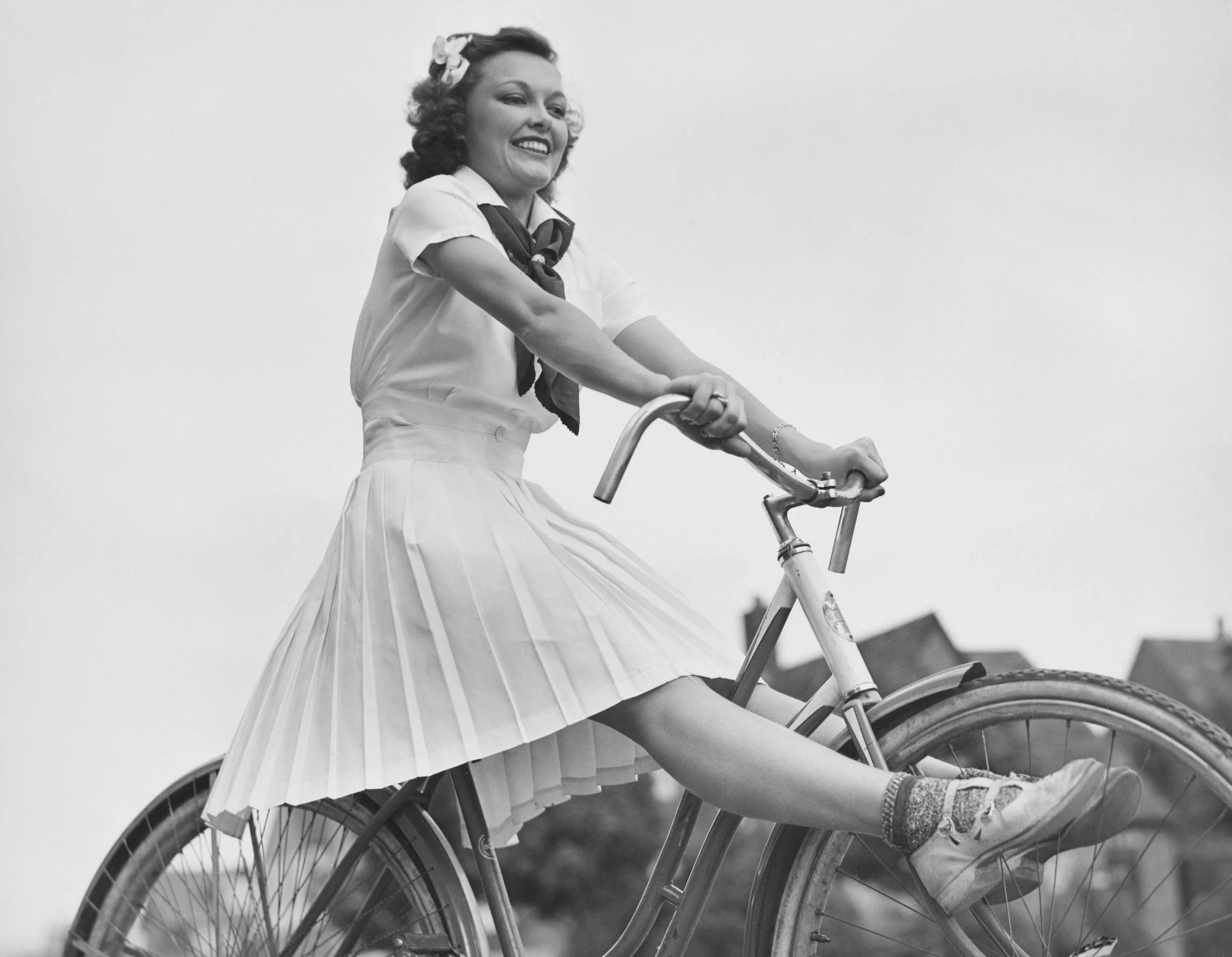 Femme souriante en période scolaire, portant une jupe plissée, un chemisier blanc et un foulard, assise sur un vélo, vue en noir et blanc.