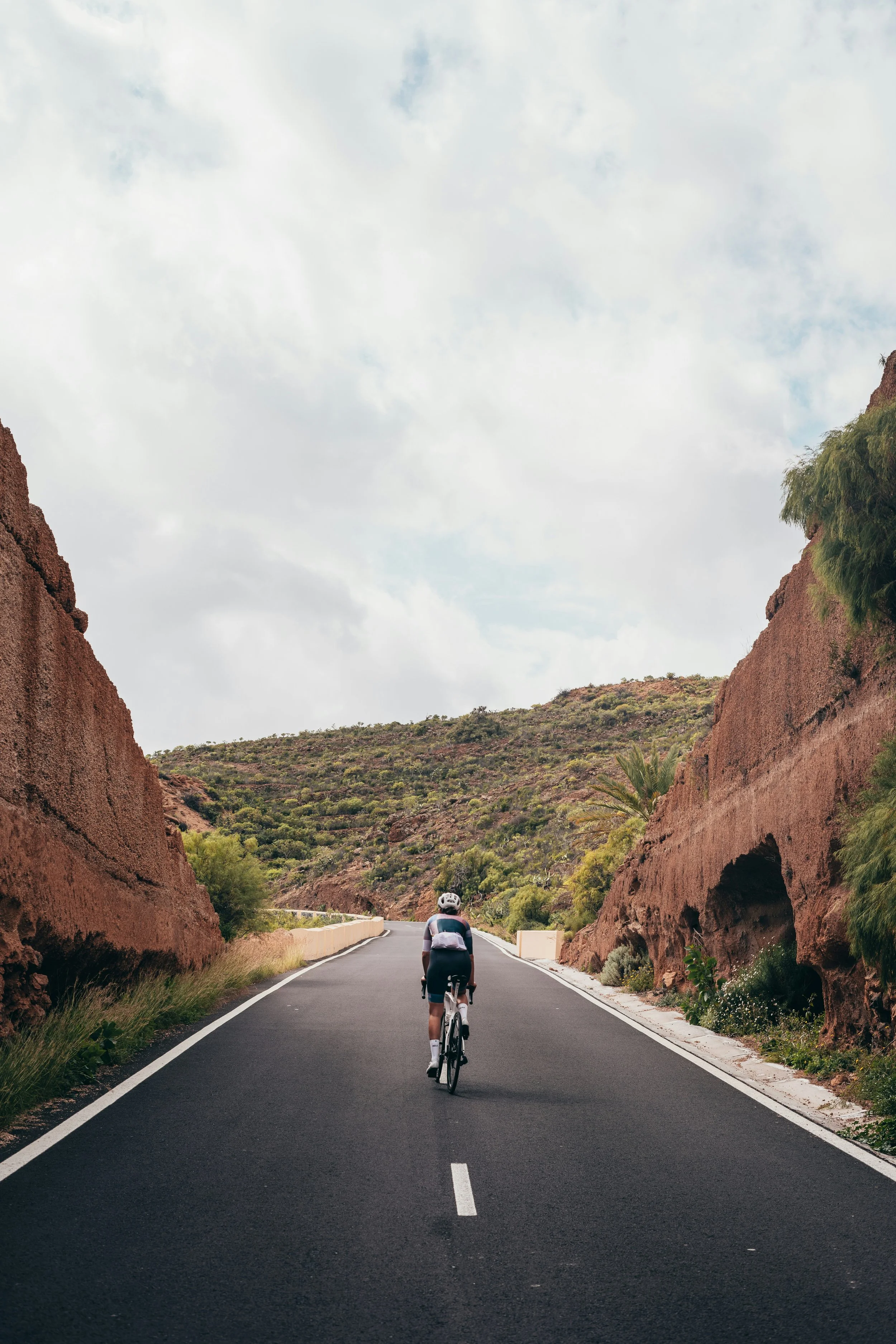Un cycliste pédale sur une route sinueuse au milieu d'un paysage de montagnes rocheuses et de végétation, sous un ciel partiellement nuageux.