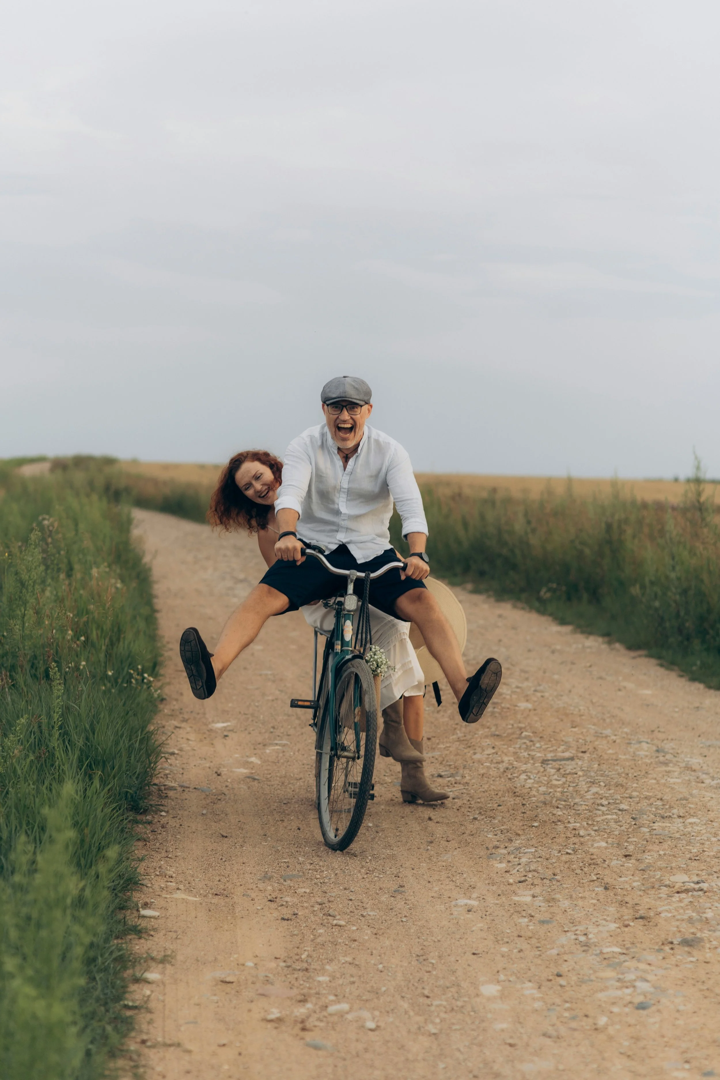 Deux personnes, un homme et une femme, sur un vélo en pleine nature sur un chemin en terre, tous deux souriants, avec un ciel nuageux.