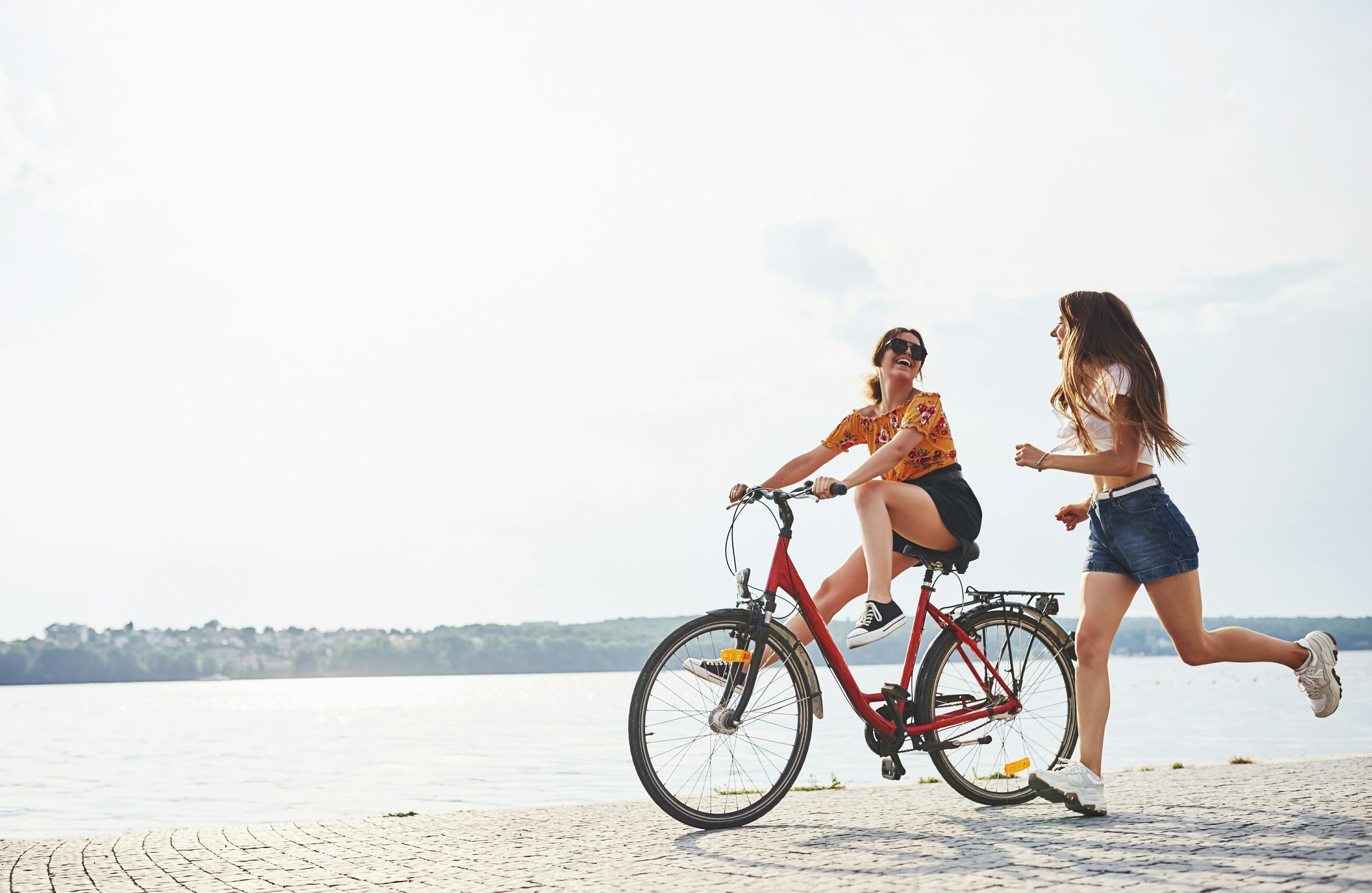 Deux jeunes femmes rient, l'une à vélo, au bord d'un lac ou d'une rivière, sous un ciel clair avec quelques nuages.