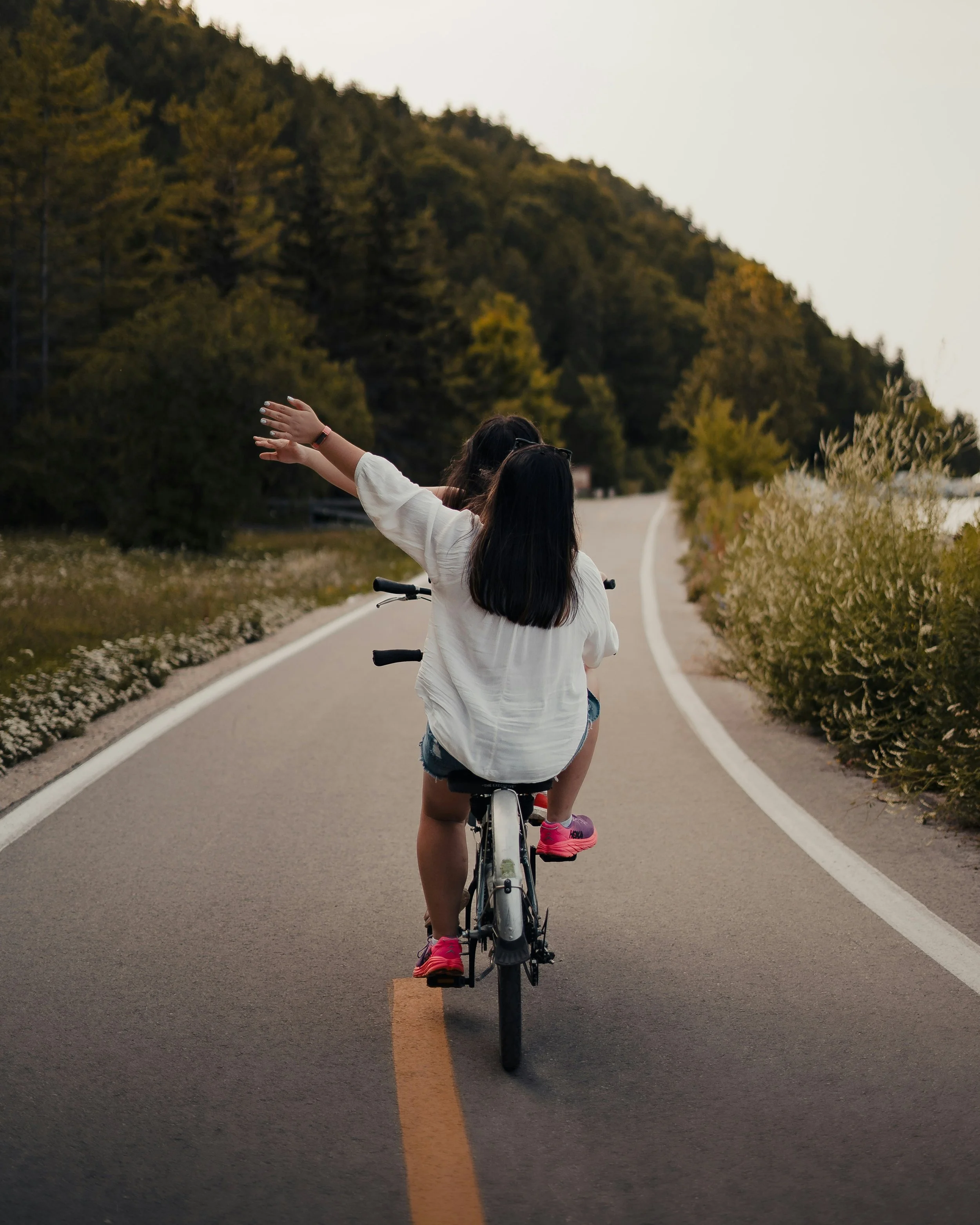 Deux jeunes filles rouillent à vélo sur une route en campagne entourée d'arbres, l'une d'elles lève le bras.