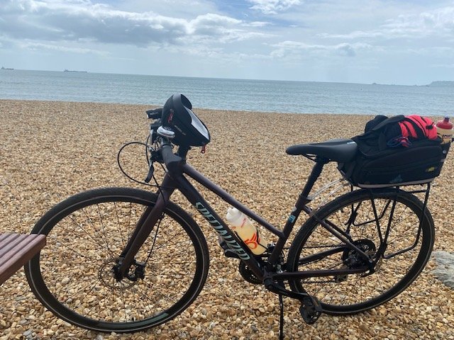 A black bicycle with a water bottle, helmet, and a bag on the rear rack parked on a pebbled beach with the ocean and cloudy sky in the background.