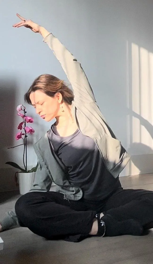 Woman practicing yoga in a seated position with one arm extended, next to a blooming orchid in a pot. Nadine McBride mat pilates in Belfast. Gadd Health and Wellness