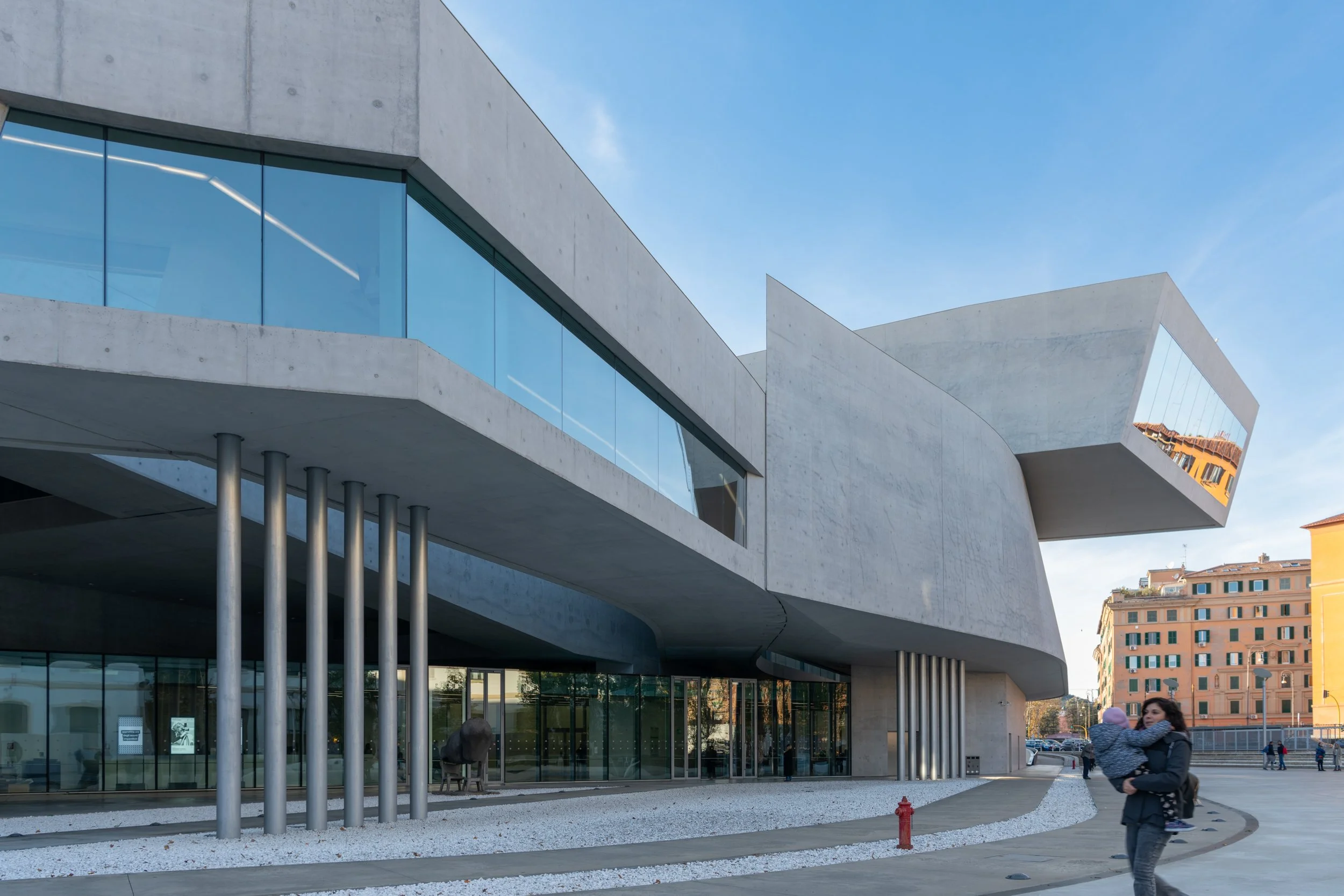 The Maxxi in Rome by Zaha Hadid Architects. A modern concrete building with large glass windows and steel support columns, with a person holding a child in the foreground and a cityscape in the background.