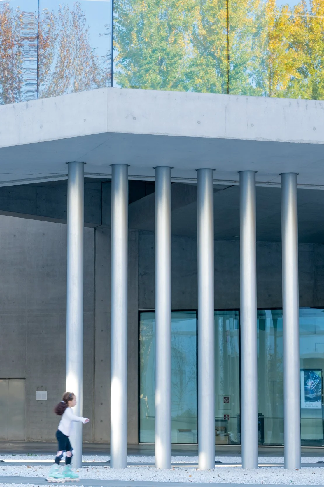 The Maxxi in Rome by Zaha Hadid Architects. A young girl rollerblading past a modern building with concrete walls and six vertical metal columns, and a glass facade reflecting trees with autumn foliage in the background.