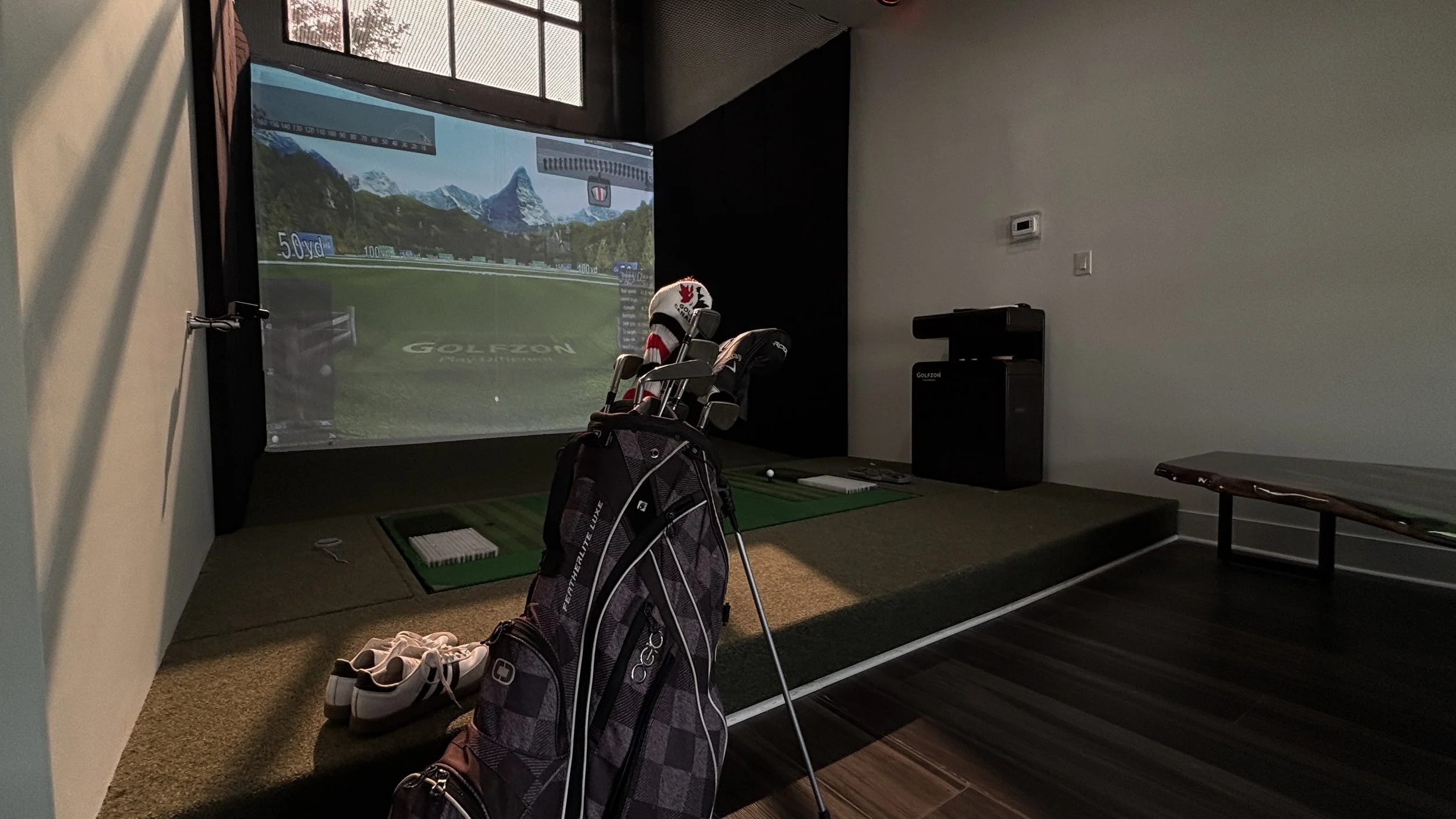 Indoor golf simulator room with a projected screen showing a golf course, a golf bag with clubs, and a pair of golf shoes on the floor.
