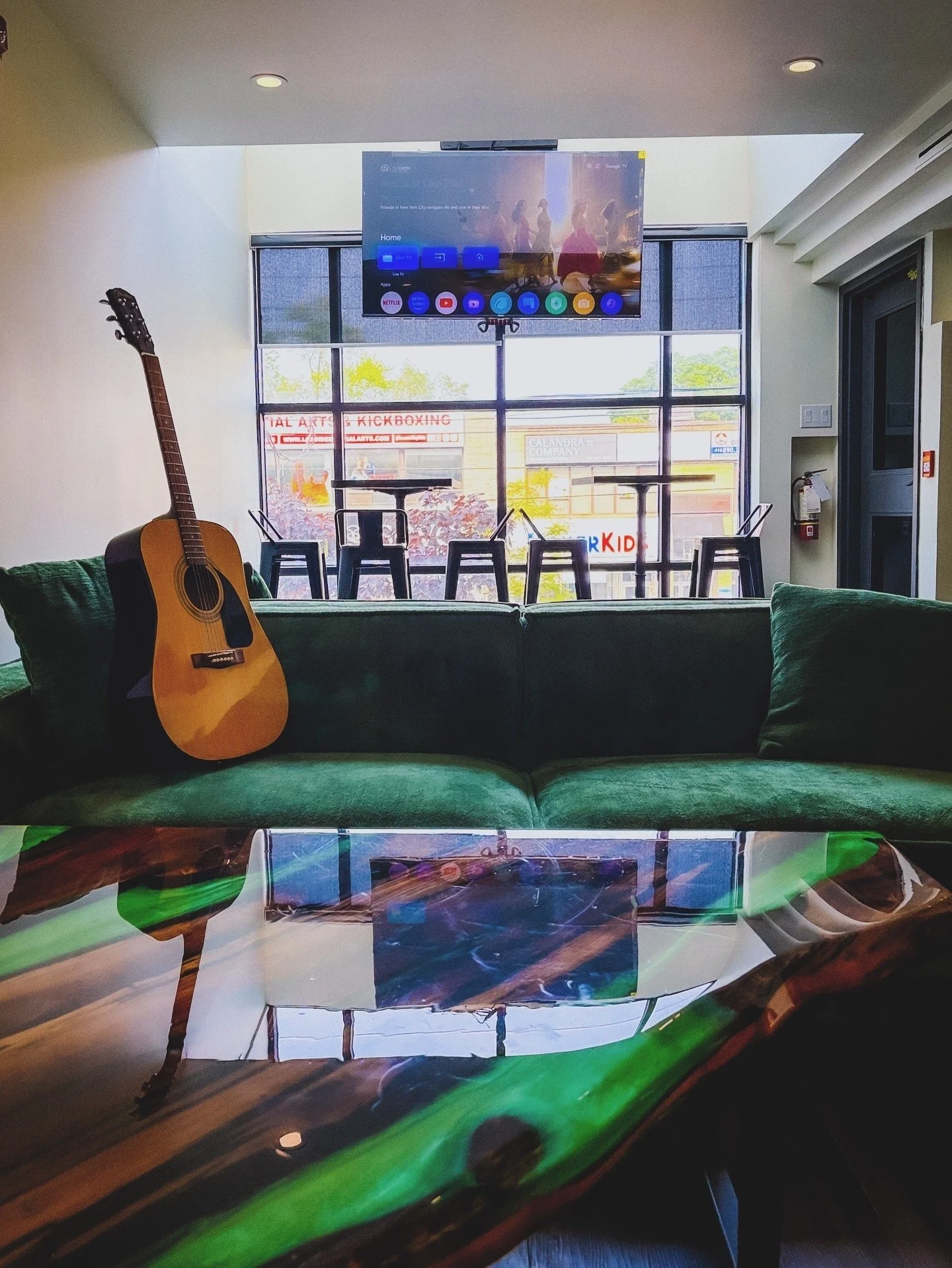 A TV mounted on the wall in a modern lounge area with a guitar resting on a green velvet couch, a colorful wooden table in the foreground, and large windows with a view of signs for a talk arts and kickboxing studio outside.