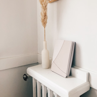 A white shelf with a white vase containing dried pampas grass and a closed book on top, against a white wall.