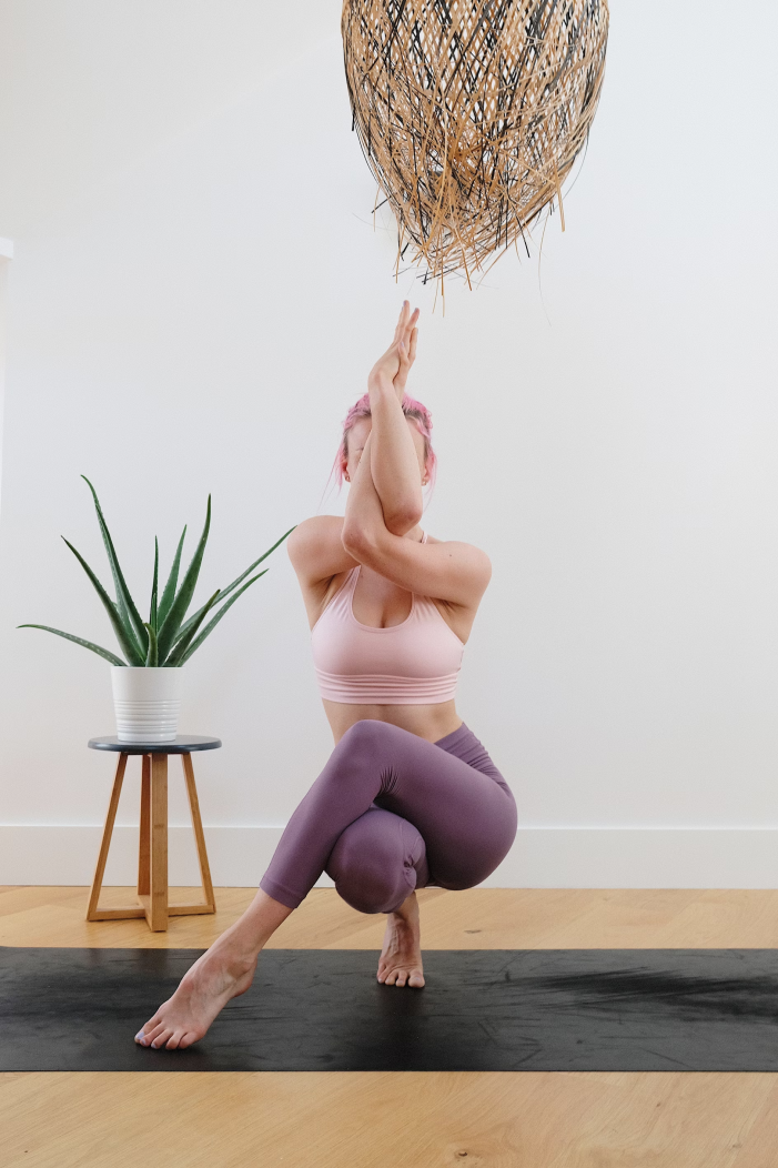 Woman practicing yoga indoors, kneeling with one knee on the ground, hands pressed together overhead, and head slightly tilted back. She is wearing a light pink sports bra and purple leggings, with a black yoga mat on a wooden floor. There is a potted plant on a small wooden stand and a large wicker light fixture hanging from the ceiling.