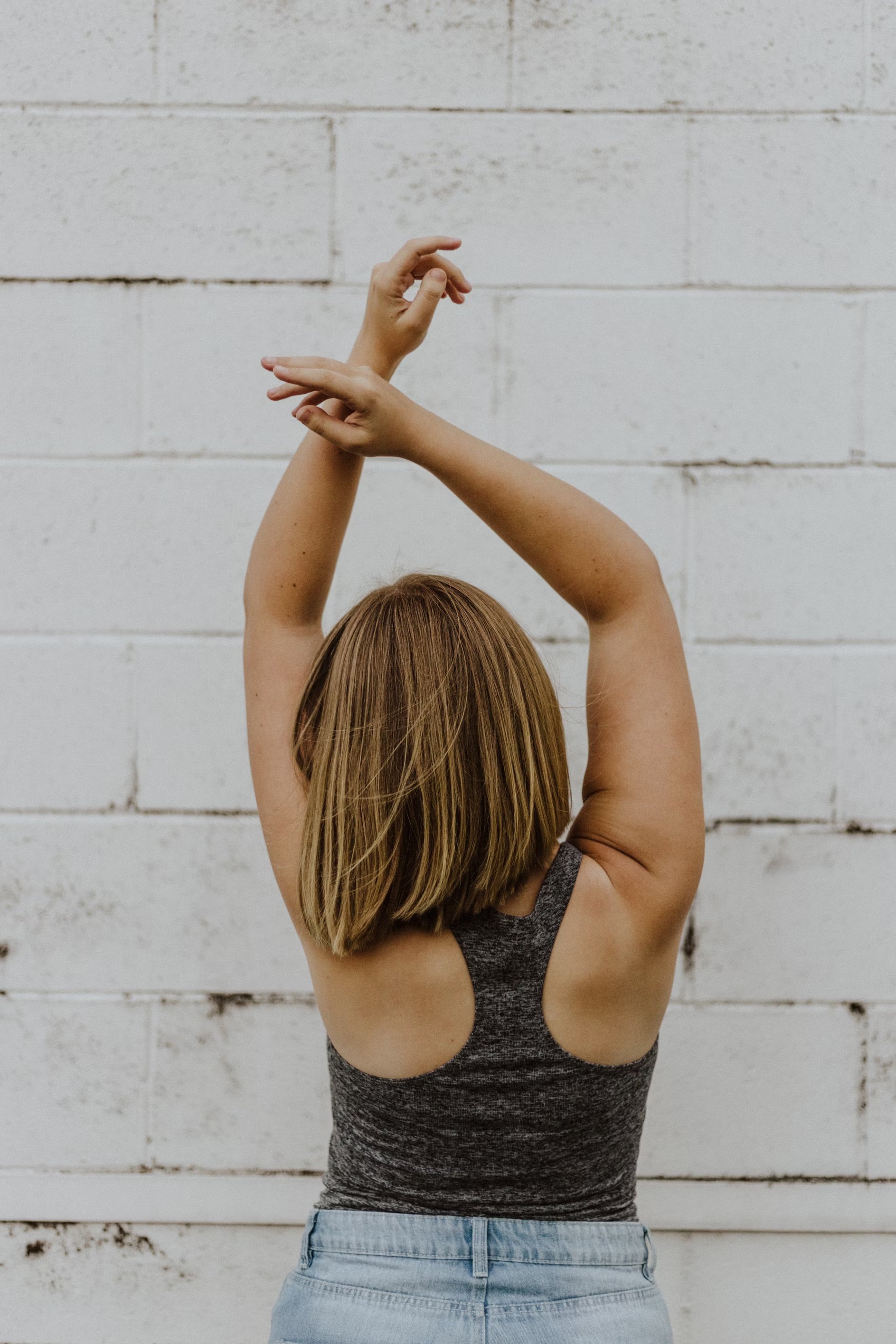 Back view of a woman with short blonde hair wearing a gray tank top and light blue jeans, posing with arms raised and crossed above her head against a white brick wall.