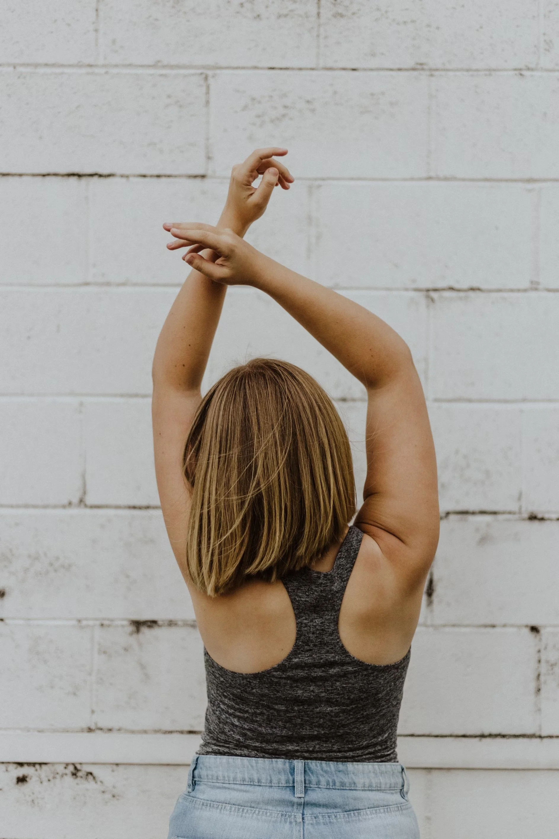 Back view of a woman with short blonde hair stretching her arms above her head against a white brick wall