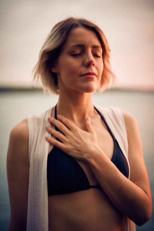 A woman with short blonde hair and closed eyes touching her neck with her hand, outdoors near water during sunset.