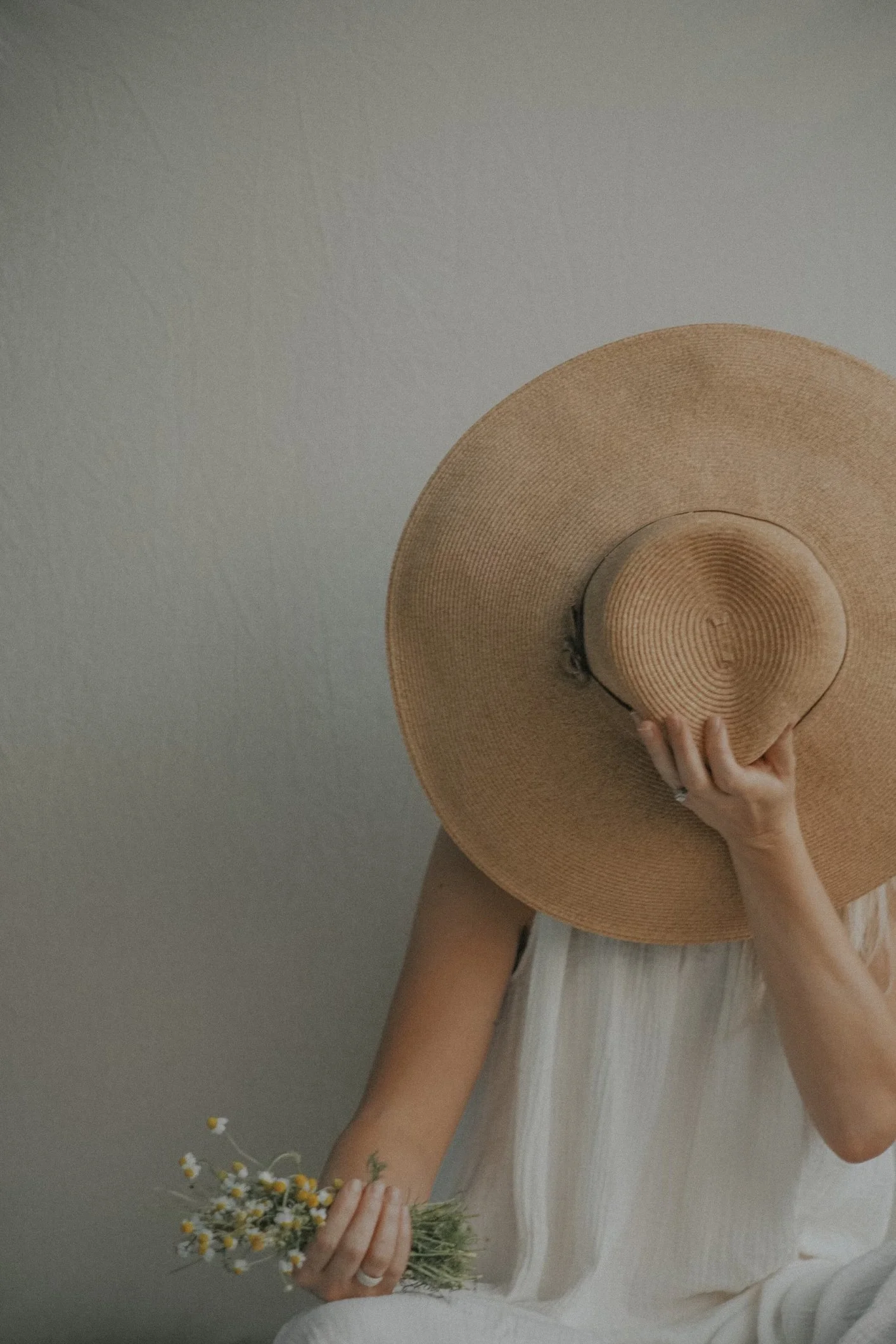 Person holding a large sun hat and a small bouquet of daisies, dressed in a light-colored sleeveless top, in front of a plain wall.